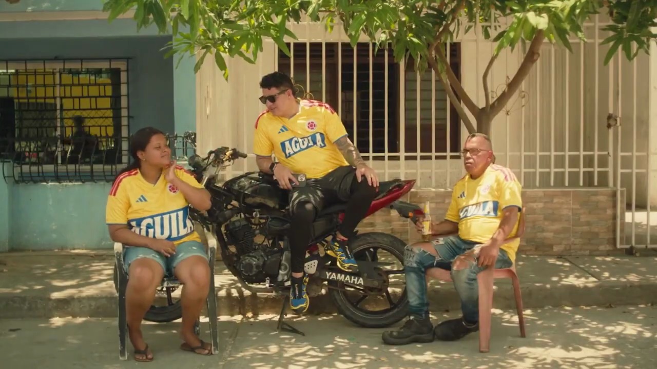 Photographie de rue diurne en plan pied de trois supporters colombiens détendus posant devant un mur bleu clair grillagé d'une maison de quartier sous un arbre, femme en maillot jaune de la sélection colombienne Adidas frappé du sponsor Aguila en gros logo blanc et bleu sur la poitrine assise sur une chaise en plastique blanche jean court bleu et tongs, homme au centre assis à califourchon sur une moto Yamaha noire à réservoir rouge en maillot jaune sponsorisé Aguila lunettes de soleil sombres pantalon en cuir noir baskets bleues et jaunes tenant un cocktail dans une gourde blanche, homme plus âgé assis à droite en maillot jaune barré Aguila jean déchiré usé et bottes noires tenant un verre orange, tronçon d'un arbre feuillu qui projette son ombre en dentelle, sol de trottoir en dalles claires, éclairage naturel chaud tropical