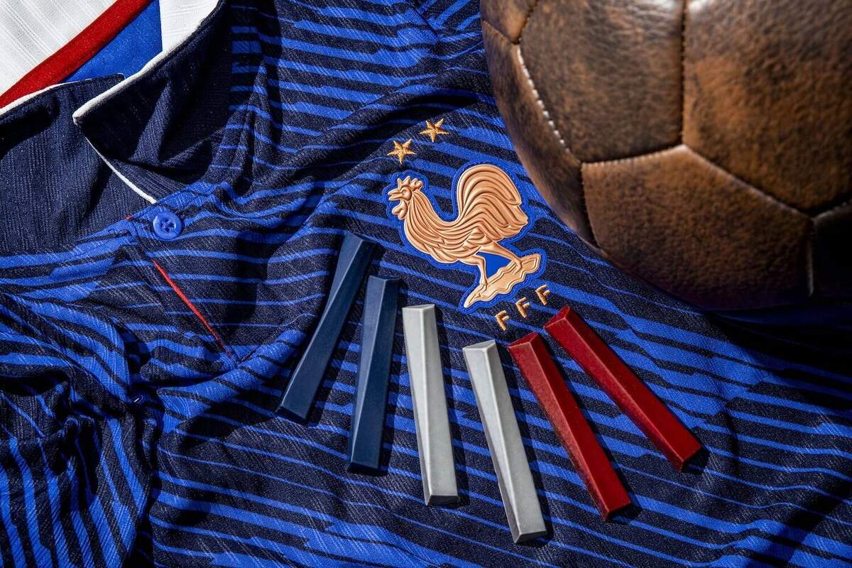Photographie macro très rapprochée en studio de la capsule Yannick Alléno x FFF dévoilée le 3 juillet 2026, cadrage serré sur un maillot officiel de l'équipe de France en jersey rayé bleu marine et bleu roi orné du coq FFF or et cuivré à trois étoiles avec col rouge blanc étroit, six sticks de chocolats pralinés couchés en éventail directement sur le maillot deux bleu marine deux gris perle et deux rouges laqués, un ballon de football vintage en cuir marron patiné cousu à l'ancienne posé en haut à droite, lumière douce et diffuse mise en scène qui suggère le rituel du soir de match, image de dossier de presse Creapills complètement différente de la couverture