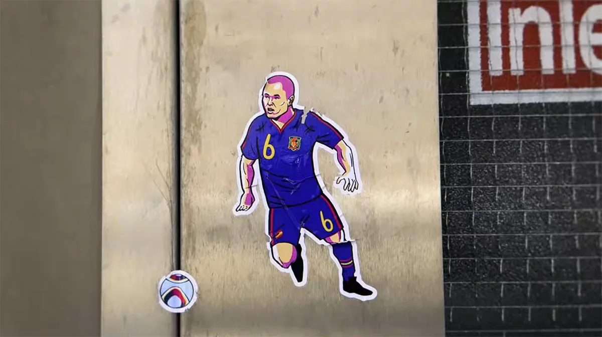 Plan extrait du film Let's Make It Iconic de BBC Creative, illustration en papier découpé d'Andrés Iniesta en maillot bordeaux numéro 6 de l'Espagne posée sur une devanture commerciale, micro-ballon collé en bas du plan, animation en stop motion réalisée par Nicos Livesey et Dan Evans, mai 2026