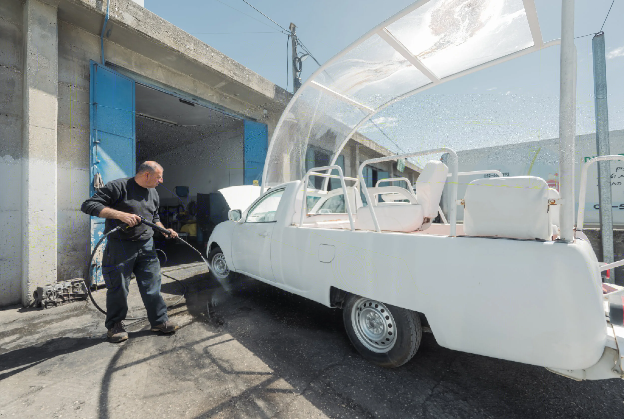 Vue dévoilée par Caritas Jérusalem le 22 juin 2026 du véhicule Vehicle of Hope dans un garage de Bethléem, ancienne papamobile blanche du pape François entièrement repeinte, plateforme arrière refermée par une carrosserie blanche pour créer une salle de soins, capot ouvert et toit transparent en arche au-dessus du véhicule, mécanicien palestinien en pull noir en train de nettoyer le pare-chocs avant au nettoyeur haute pression