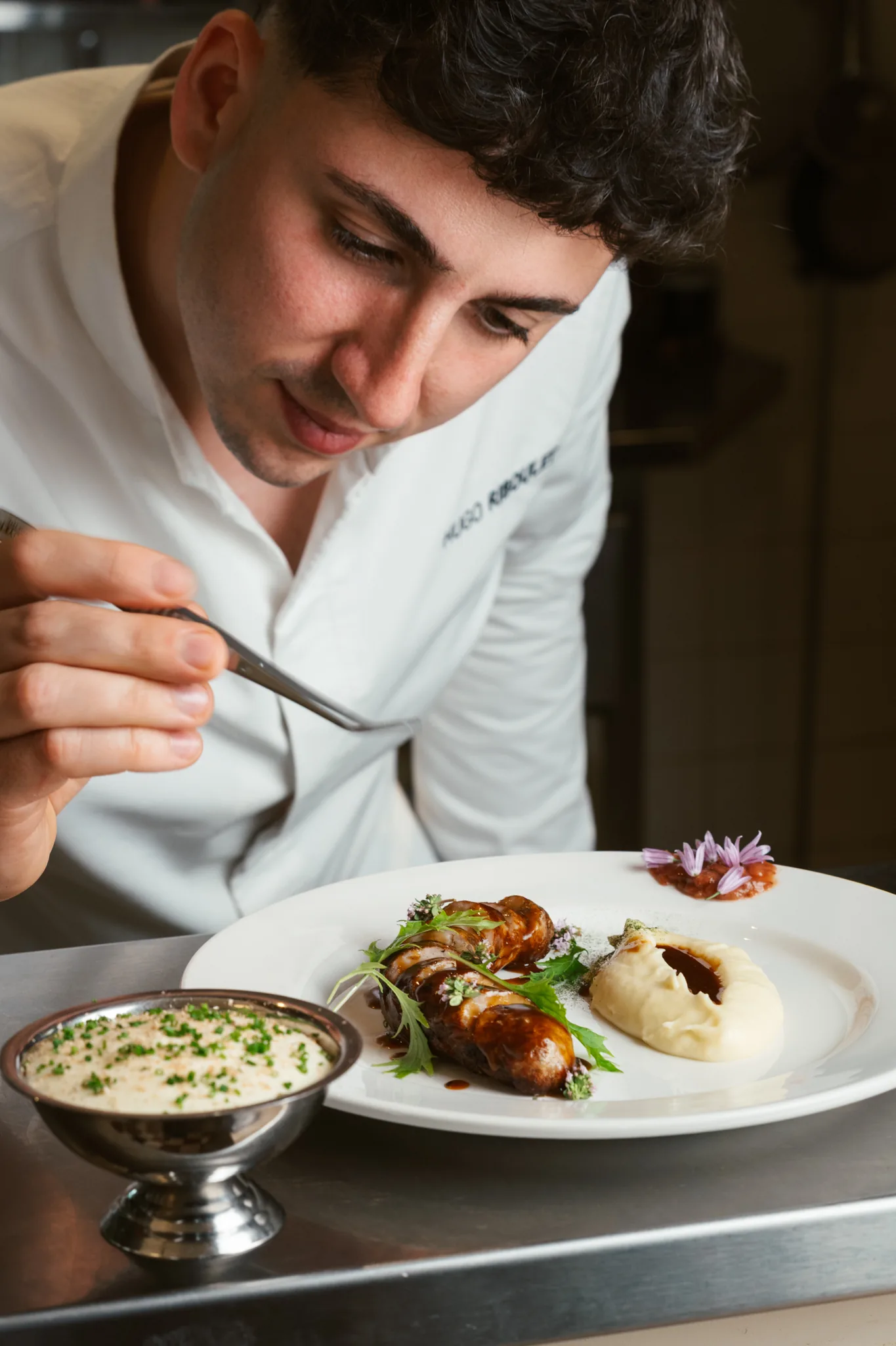 Le chef Hugo Riboulet en train de dresser une assiette gastronomique en cuisine du restaurant Le Cinquante, gros plan sur les gestes de précision, assiette composée d'une viande nappée de sauce brune, purée et fleur comestible, opération Carrefour x BeInfluence mai 2026