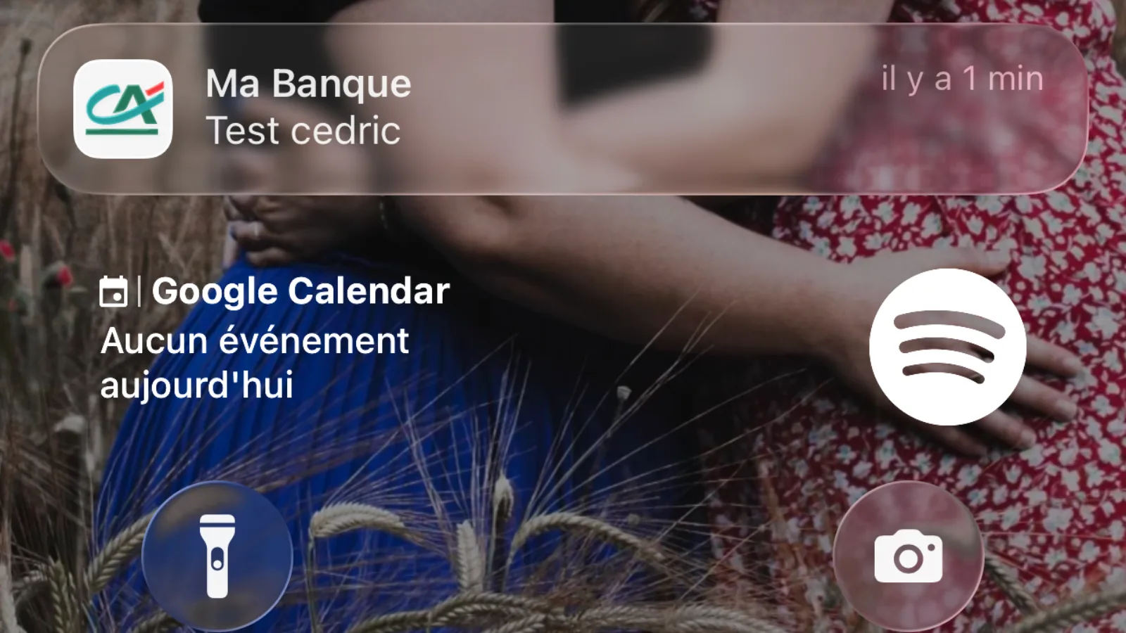 Écran de verrouillage d'iPhone le 9 juin 2026 montrant la notification Ma Banque Test cedric reçue par un client du Crédit Agricole, fond d'écran d'un couple enlacé dans un champ de blé au coucher du soleil, widgets Google Calendar et Spotify visibles en dessous, raccourcis lampe torche et appareil photo en bas, image diffusée par Clubic
