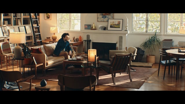Photogramme du film How Can I Communicate Better With My Mom? signé Mother London et réalisé par Jeff Low via Biscuit Filmworks pour Anthropic Claude, plan large d'un grand salon californien aux baies vitrées, sol bois clair, canapé en cuir caramel, bibliothèque haute, lampe en céramique au premier plan, cheminée en marbre blanc, fauteuils mid century en bois et chaises tournées vers le centre, un homme assis sur le canapé en jean et veste sombre tient un mug et écoute une interlocutrice hors champ, lumière dorée d'après-midi