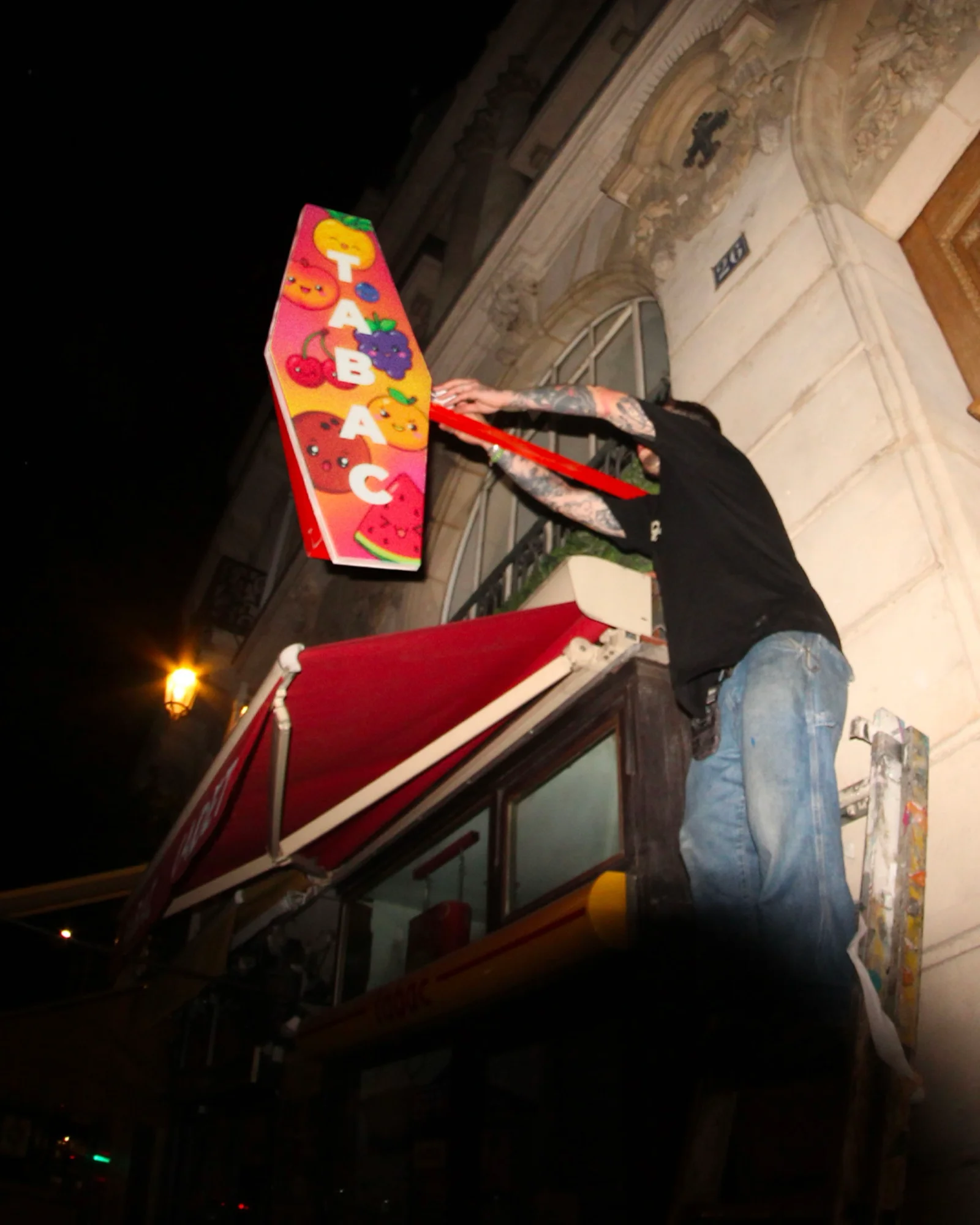 Photo prise en contre-plongée en pleine nuit dans une rue parisienne pendant l'installation guerilla du 19 au 20 juin 2026 pour la campagne Contre-Feu signée Buzzman, un colleur tatoué en tee-shirt noir et jean bleu perché au-dessus d'un store rouge de bureau de tabac fixe au ruban adhésif rouge une carotte pop détournée aux couleurs kawaï rose orange et jaune imprimée de fruits souriants cerise raisin orange pastèque et lettrage T A B A C en verticale blanc, façade haussmannienne pierre claire lampadaire allumé au deuxième plan ciel noir, cliché voleur qui montre le geste d'installation loin du plan panoramique de la campagne officielle