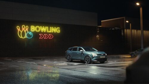 Photogramme officiel du film de lancement européen du nouveau Dacia Striker signé Publicis Conseil et réalisé par Cole Webley pour BIG Productions dévoilé le 8 juillet 2026, plan large nuit sur un parking de bowling américain, mur de brique brune barré à gauche par une enseigne néon aux couleurs vives quilles blanches boule de bowling bleu clair et lettrage jaune BOWLING flanqué de trois chevrons rouges pointant vers la droite, au centre-droit le nouveau crossover Dacia Striker gris bleu métallisé profil trois-quarts avant capot plat feux étroits jantes noires garé sous un lampadaire, sol goudronné humide reflétant les néons rose et bleu et bordé de mur en tôle nervurée en arrière-plan