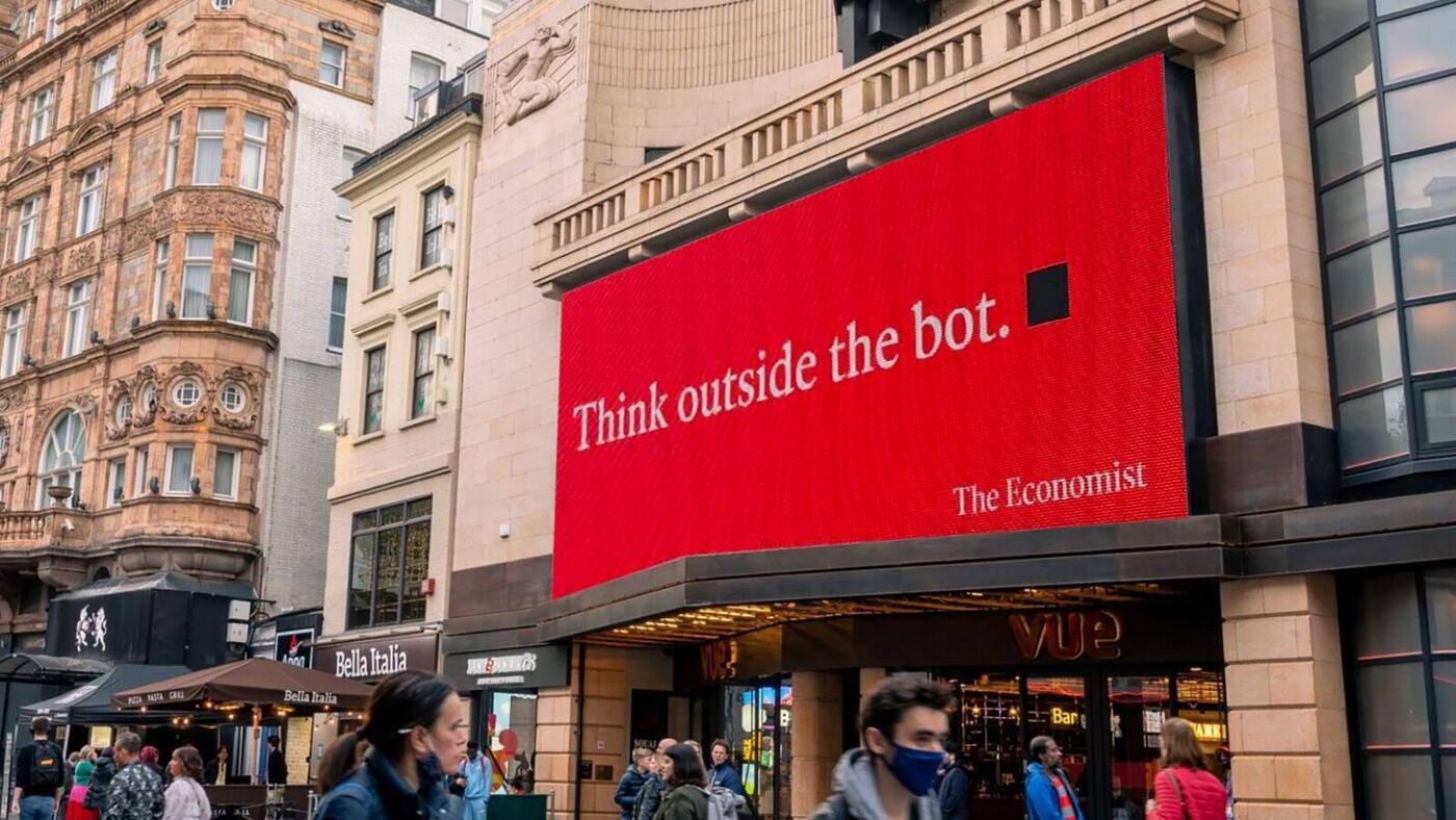Photographie en plan large diurne à hauteur de rue de la nouvelle campagne d'affichage OOH The Economist White out of Red signée Cocogun installée à Leicester Square à Londres début juillet 2026, immense écran DOOH rouge sang encastré dans la façade art déco pierre claire du cinéma Vue avec balcons balustrés et bas-reliefs de danseurs, texte en typographie Roman blanche « Think outside the bot » suivi du petit carré noir emblématique et de la signature The Economist en bas à droite, façades voisines de la brasserie Bella Italia et d'un immeuble haussmannien clair, terrasse avec parasol au sol, passants indifférents en manteau et casquette rouge en bas de cadre, ciel gris pommelé au-dessus de la balustrade