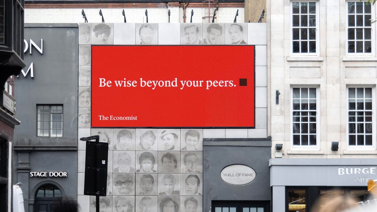 Photographie plan large diurne d'une seconde affiche de la saga The Economist White out of Red signée Cocogun début juillet 2026, immense écran DOOH horizontal rouge sang incrusté sur une façade Wall of Fame en carrelage crème parsemée de portraits noir et blanc de célébrités britanniques comiques, texte blanc en gros à empattements « Be wise beyond your peers » suivi du carré noir signature et de la mention The Economist en bas à gauche, façade voisine d'un pub Stage Door à l'auvent noir et d'un Burger à droite, ambiance de rue londonienne calme peu passante, ciel gris blanc, image nettement différente de la scène de Leicester Square utilisée en couverture