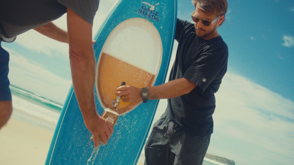 Séquence tirée du film de lancement du Beerboard réalisé par Lauchlan Dempsey pour Gage Roads Brew Co le 30 juin 2026 sur une plage de Perth, plan rapproché en contre-plongée d'un jeune homme blond barbu vêtu d'un tee-shirt noir et de lunettes de soleil miroir tenant à la verticale la planche de surf bleue turquoise dont la chambre transparente laisse couler la Summer Ale ambrée depuis le robinet cuivré fixé sur la board dans un verre pinte tenu par une seconde main hors cadre, mer et ciel filés en arrière-plan