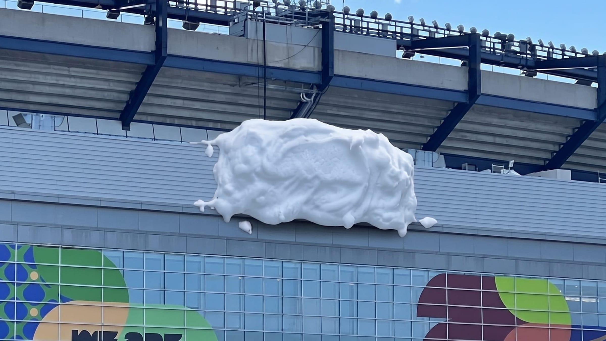 Vue aérienne du Gillette Stadium dévoilée le 21 juin 2026 pendant la Coupe du Monde 2026, terrain de football vert vif vu du ciel entouré de 64 146 sièges chacun recouvert d'un sticker bleu imprimé en faux relief de mousse à raser blanche dégoulinante, l'effet global créant des arabesques crémeuses sur les tribunes vu de très haut, ciel bleu pâle de fin d'après-midi à Foxborough Massachusetts