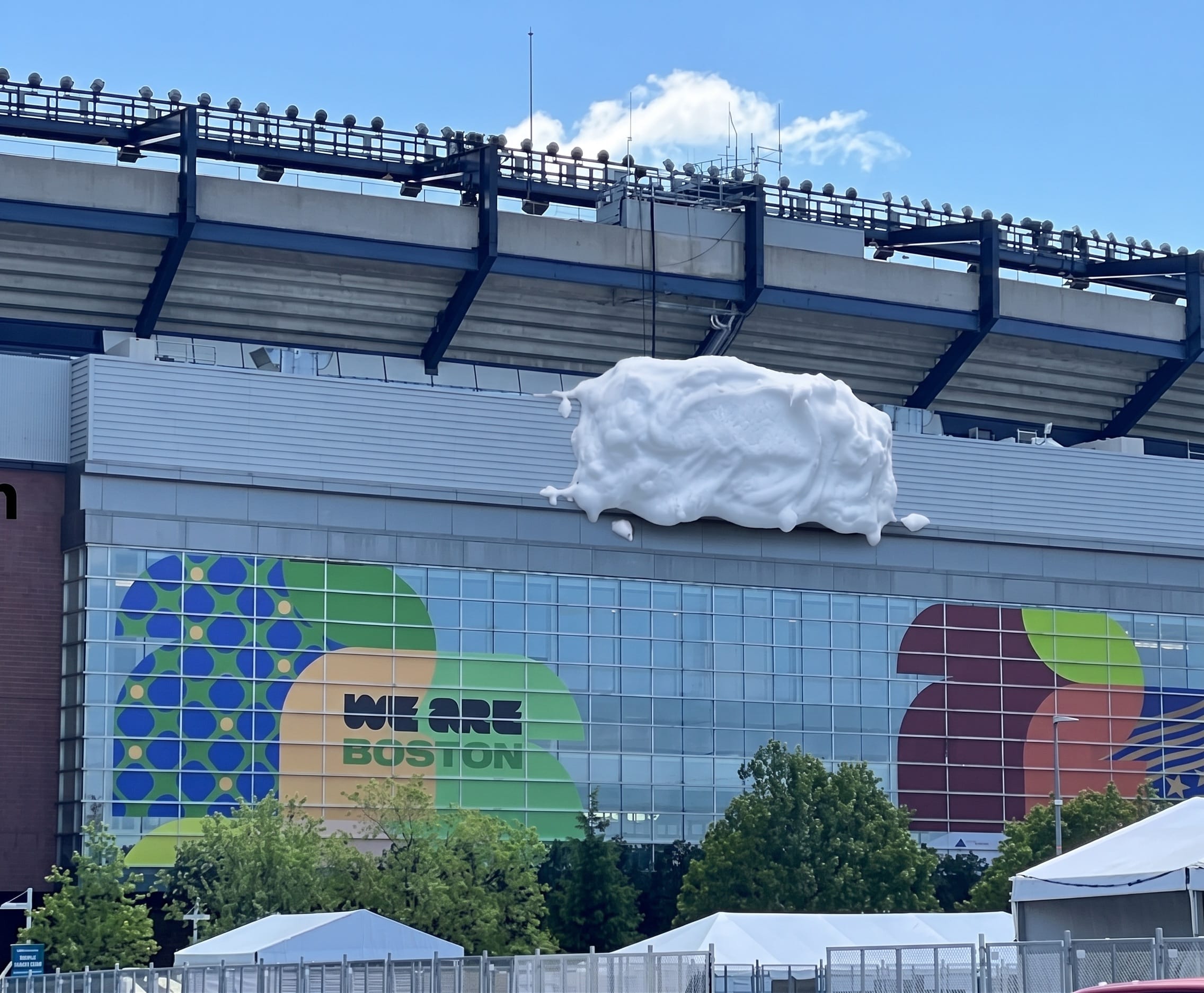 Gros plan dévoilé le 21 juin 2026 sur un siège du Boston Stadium recouvert du sticker Gillette mousse à raser, sticker en relief mat imitant la texture moussue d'une crème à raser fraîche avec gouttes et bulles tridimensionnelles peintes en blanc crémeux sur fond bleu Gillette, vis et coutures du fauteuil visibles en arrière-plan flou, environnement de stade avec rampe et marchepied