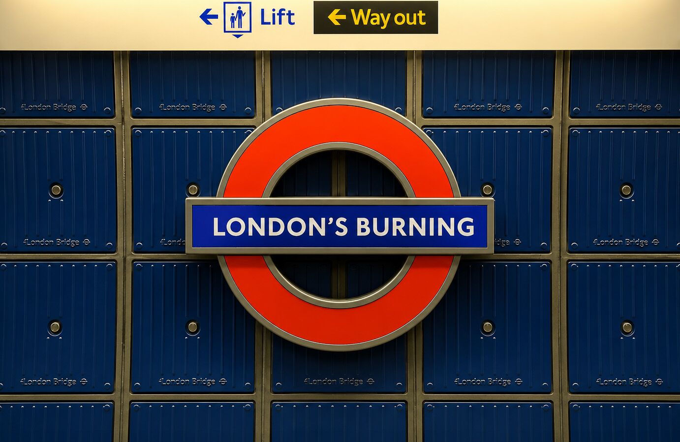 Photographie serrée prise à hauteur d'homme dans un couloir de la station londonienne London Bridge pendant l'opération guerilla de Greenpeace UK de fin juin 2026, panneau officiel de la London Underground bombé rond avec cercle rouge et barre bleue sur laquelle Greenpeace a collé le texte London's Burning en typographie sans serif blanche à la place de London Bridge, fond de mur mosaïqué en carreaux bleu marine verticaux gravés du toponyme London Bridge en discret, deux panneaux jaunes signalétiques Lift et Way out en tête de cadre avec pictogramme personne debout, éclairage plat et frontal, tirage documentaire brut