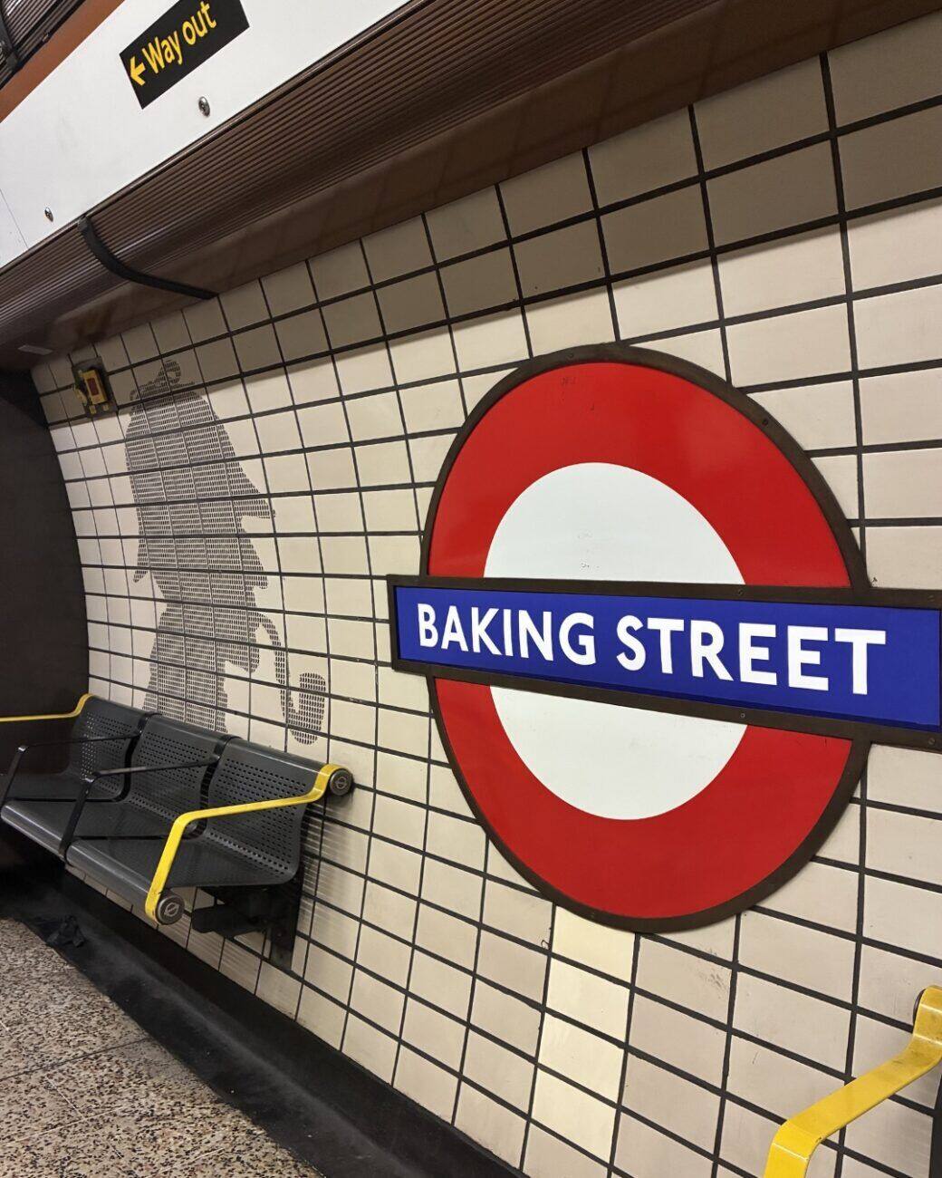Photographie prise en plan large à Baker Street pendant la campagne Greenpeace UK sur les stations rebaptisées du métro londonien, panneau rond London Underground à cercle rouge et barre bleue collé du nouveau nom BAKING STREET en typographie blanche caractères stencil serrés, en fond mur en carrelage blanc à joints noirs typique de la station Baker Street ligne Bakerloo silhouette pochoir de Sherlock Holmes en gris à gauche du panneau, banc gris et jaune vide en amorce en bas à gauche, cadrage frontal et vide de voyageurs qui montre le détournement de manière calme et documentaire, scène complètement différente du plan London Bridge en cover