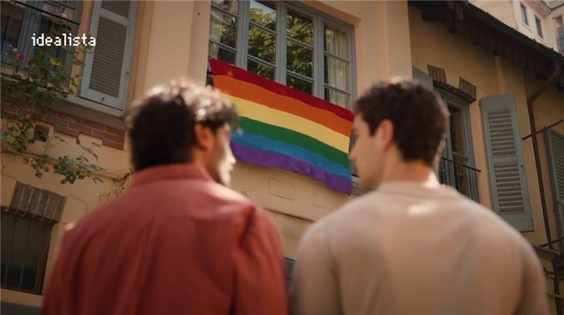 Photogramme du spot Ridicoli! signé Gibbo&Lori pour Idealista, vue de dos rapprochée des deux jeunes hommes du couple en chemise rouge orangée et pull beige observant ensemble depuis la cour de l'immeuble milanais le grand drapeau arc-en-ciel fait au crochet à mailles serrées accroché en façade au balcon du premier étage de la maison jaune crème aux volets bleus, logo Idealista pixel blanc en haut à gauche