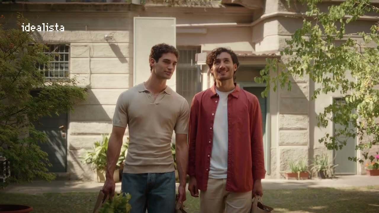 Photogramme du spot Ridicoli! signé Gibbo&Lori pour Idealista produit par The Big Mama et diffusé en Italie du 21 au 27 juin 2026 pendant le Pride Month, plan large d'une cour d'immeuble milanais en pierre claire bordée d'arbres ombrageux, au centre deux jeunes hommes souriants en chemise polo beige et chemise rouge orangée portant chacun un sac de courses en kraft avec des légumes verts, lumière chaude de fin d'après-midi, logo Idealista en pixel art blanc en haut à gauche