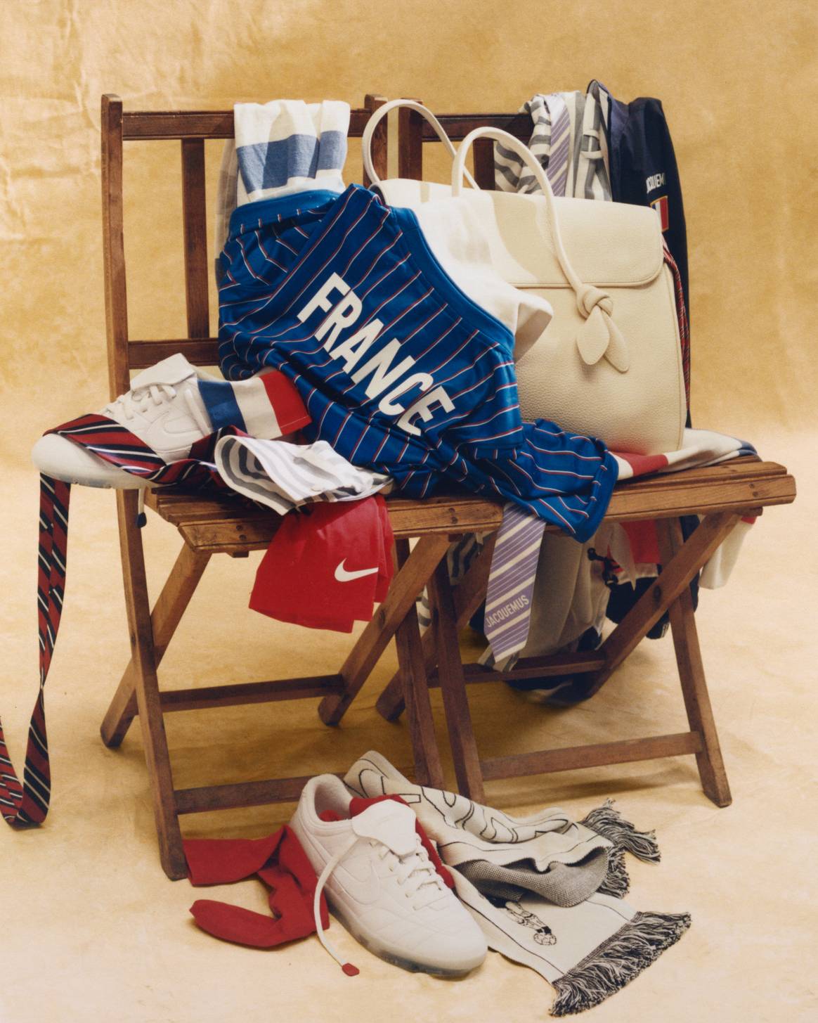 Nature morte officielle de la collection Jacquemus x Nike x FFF posée sur une chaise en bois, maillot bleu FRANCE plié, sac tote, sneakers blanches, écharpe et accessoires aux couleurs des Bleus, photographiée pour le lancement du 11 juin 2026 et diffusée par Fashion United