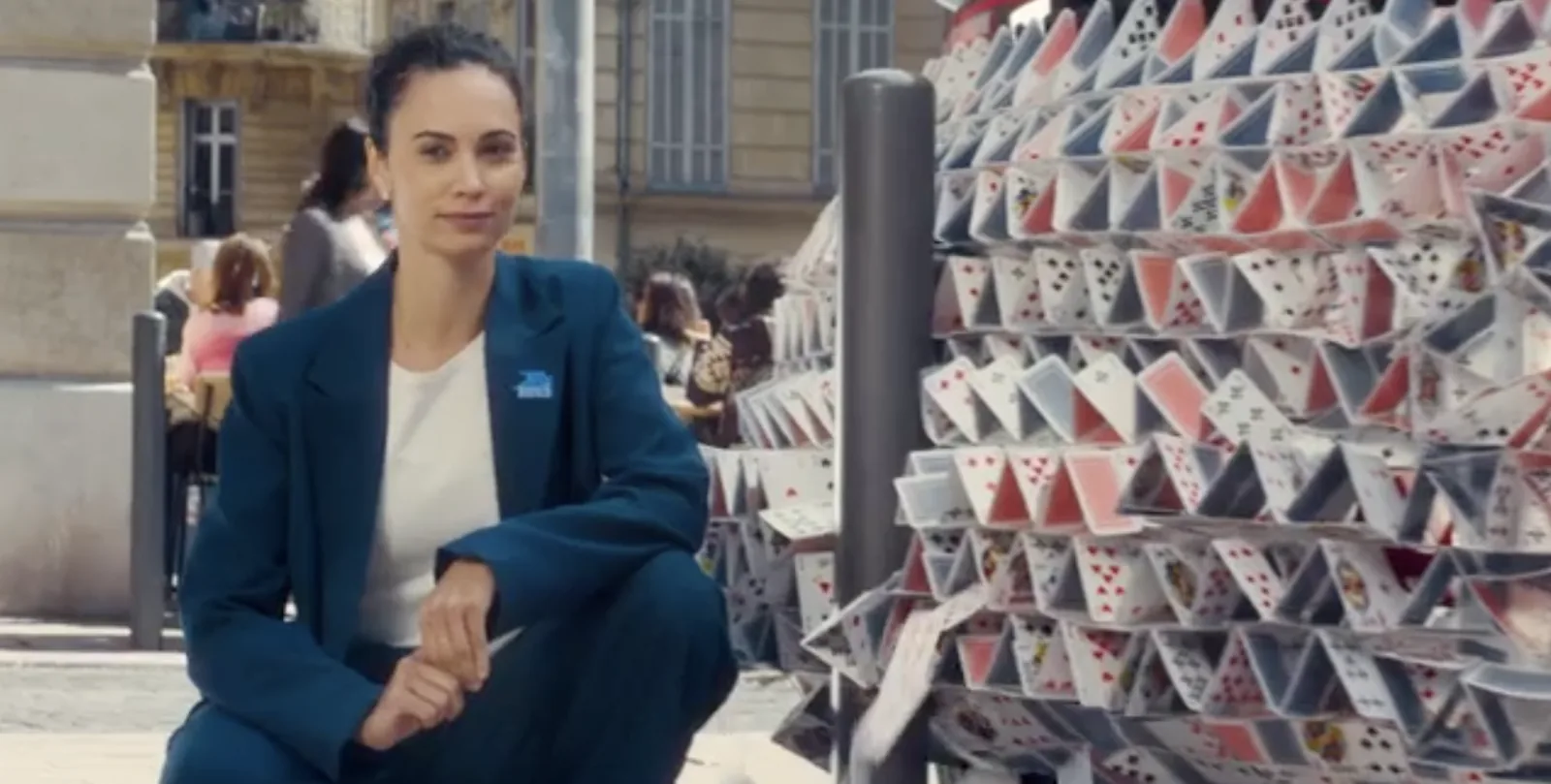 Plan d'une scène extérieure du film Avoir un temps d'avance de La Banque Postale tourné à Marseille en mai 2026, une conseillère bancaire en tailleur bleu assise devant un immense château de cartes installé en plein milieu d'une rue marseillaise, terrasse de café et passants visibles à l'arrière, tournage réel avec maquettes à taille humaine avant les retouches 3D