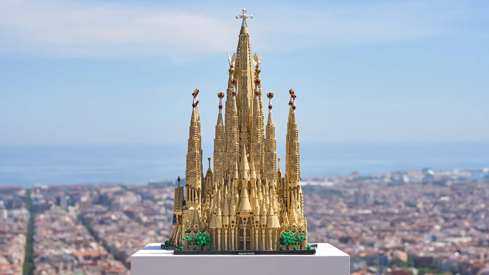 LEGO Architecture Sagrada Família dévoilé le 4 juin 2026 pour le centenaire de la mort d'Antoni Gaudí, vue de la maquette en briques beiges posée sur un socle blanc en hauteur, avec en arrière-plan le panorama de Barcelone et la mer Méditerranée, set numéro 21065 conçu par Rok Žgalin Kobe