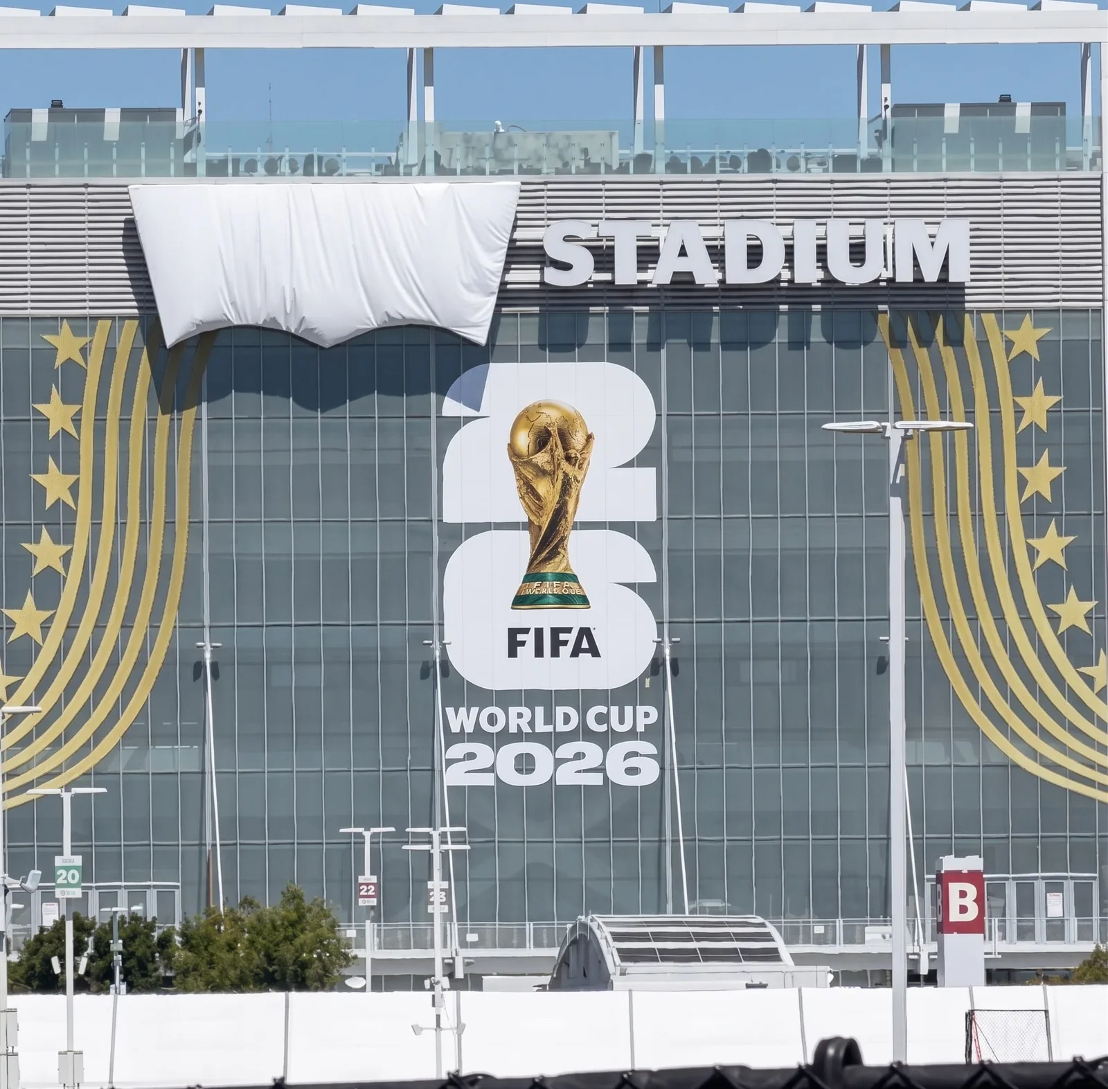 Façade vitrée du Levi's Stadium en Californie le 13 juin 2026 pendant la Coupe du Monde 2026, le logo Levi's batwing est dissimulé sous une grande bâche blanche froissée pour respecter la politique clean stadium imposée par la FIFA, panneau STADIUM en lettres rouges et logo officiel FIFA World Cup 2026 visibles à côté de la bâche, la silhouette du logo reste néanmoins parfaitement identifiable