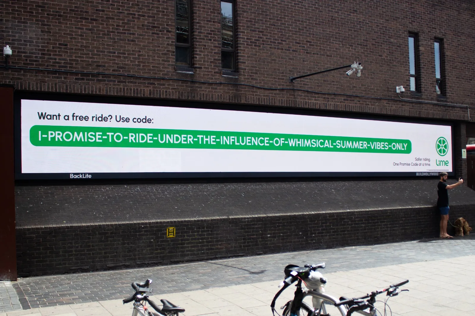 Affichage géant Lime de la campagne Promise Codes lancée le 2 juin 2026 à Londres par l'agence The Or, devanture en brique rouge avec un panneau OOH numérique BackLite, code promo géant à recopier dans l'application Lime I-PROMISE-TO-RIDE-UNDER-THE-INFLUENCE-OF-WHIMSICAL-SUMMER-VIBES-ONLY avec vélos électriques verts garés au premier plan