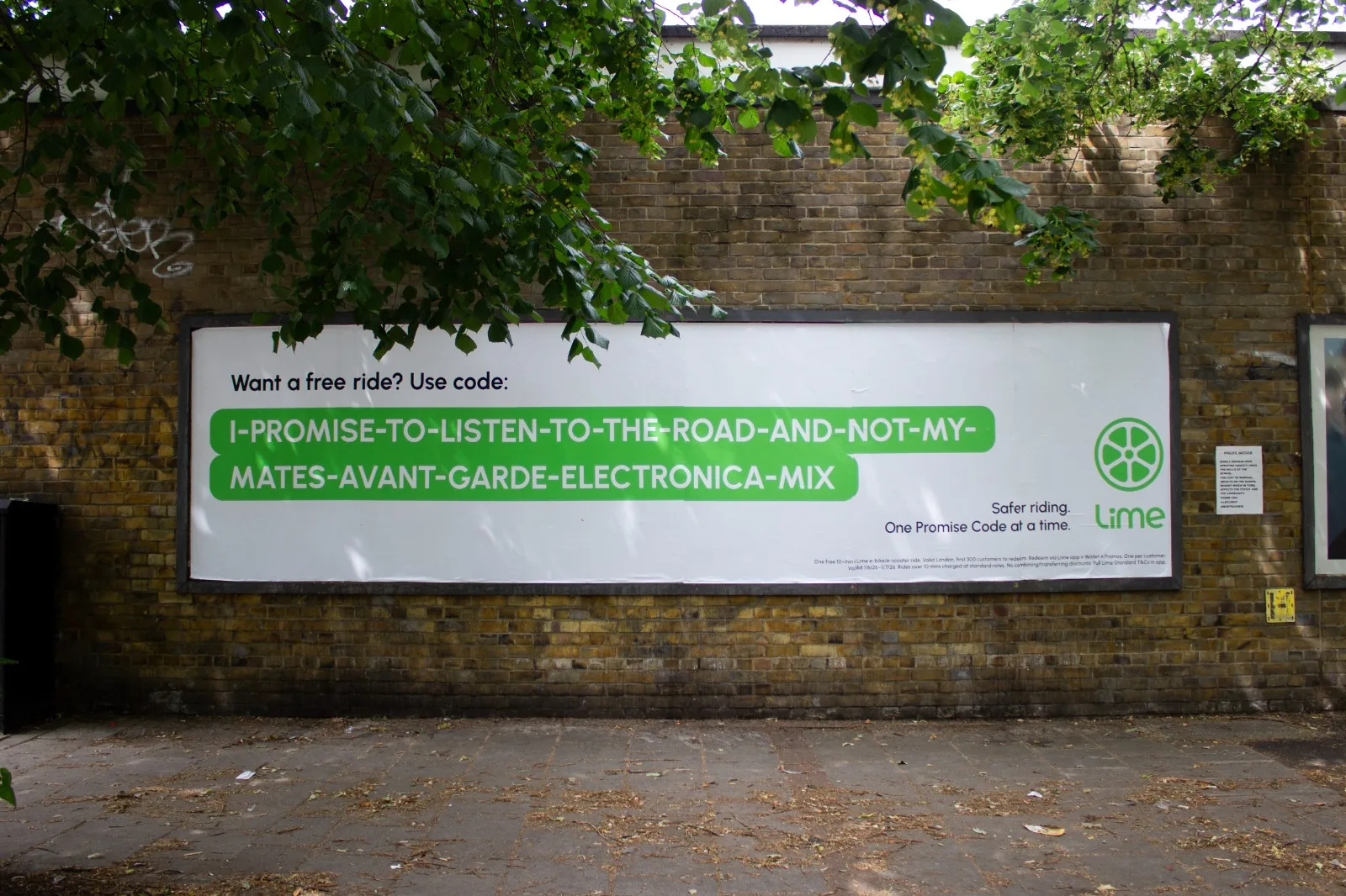 Affichage Lime Promise Codes placardé sur un mur de brique jaune à Londres encadré de feuillages d'arbres, deuxième visuel de la campagne signée The Or lancée le 2 juin 2026, code promo géant I-PROMISE-TO-LISTEN-TO-THE-ROAD-AND-NOT-MY-MATES-AVANT-GARDE-ELECTRONICA-MIX en vert Lime sur fond blanc avec slogan Safer riding, One Promise Code at a time