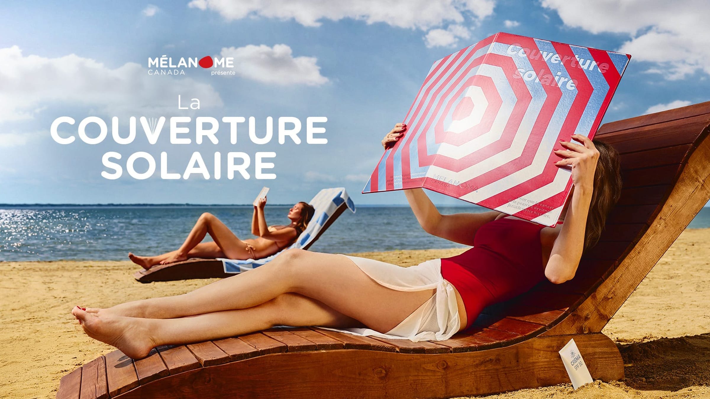 Photographie hero de campagne diurne en plan pied panoramique de la campagne Melanoma Canada présente La Couverture Solaire signée Rethink lancée le 8 juillet 2026, cadrage horizontal sur une plage de sable clair face à l'océan calme scintillant, au premier plan à droite une femme brune allongée en maillot une-pièce rouge écarlate sur un transat bois foncé qui tient à deux mains devant son visage une grande couverture de livre géante rectangulaire au motif de parasol vue du dessus en cercles concentriques rouges blancs et bleus avec la mention Couverture Solaire, au second plan à gauche une deuxième femme brune allongée en bikini beige sur un transat lit un livre au format classique sans protection, ciel légèrement nuageux bleu et blanc, gros logo blanc MÉLANOME CANADA présente et intitulé LA COUVERTURE SOLAIRE en typographie serif blanche à gauche du visuel, sable clair, éclairage naturel solaire chaud