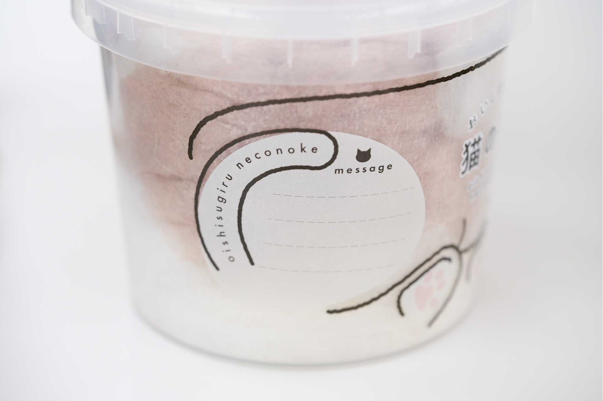 Gros plan sur le packaging du pot Necoichi oishisugiru neconoke photographié le 17 juin 2026, contenant cylindrique en plastique transparent avec barbe à papa rosée et grise à l'intérieur, étiquette illustrée d'une silhouette de chat dessinée au trait noir et d'une zone message blanche avec lignes pointillées pour écrire à la main, lettrage horizontal oishisugiru neconoke et pictogramme tête de chat avec mention message, fond blanc neutre