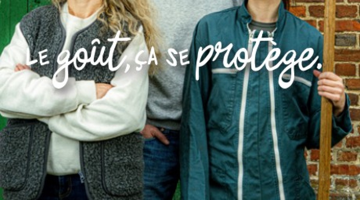 Visuel principal de la campagne d'affichage Le goût ça se protège signée Asgard pour les Fromages AOP de Normandie dévoilée le 17 juin 2026, deux portraits cadrés au buste d'éleveurs normands côte à côte devant un mur de briques rouges et un volet vert, l'une porte un gilet en laine grise sur un pull beige et croise les bras, l'autre porte une veste de travail vert sapin et tient une fourche en bois, signature manuscrite blanche LE GOÛT ÇA SE PROTÈGE incrustée en surimpression sur les vêtements