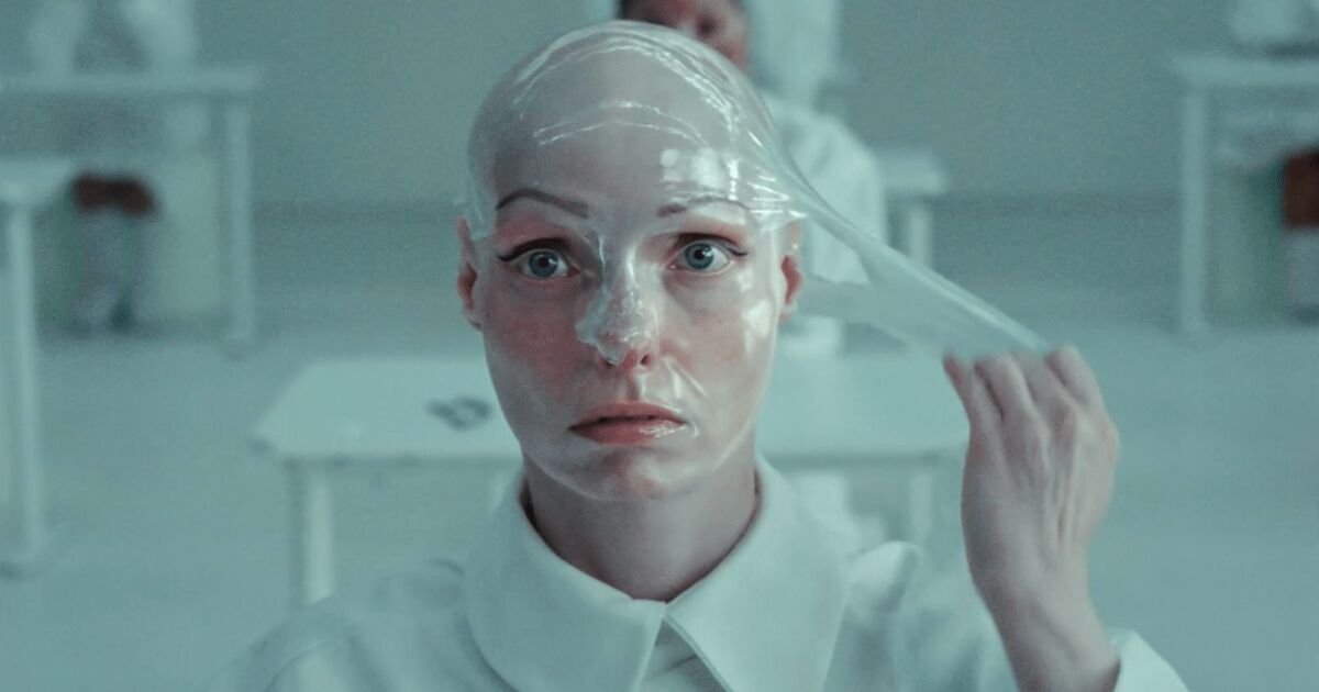 Photogramme du film The Periodic Fable signé Uncommon Creative Studio pour The Ordinary réalisé par Olivia De Camps, plan rapproché d'une femme assise dans un environnement clinique blanc, visage entièrement recouvert d'un masque de soin blanc crémeux uniforme épais qui dissimule cheveux et oreilles, main droite tenant un outil de massage facial posé sur la tempe, silhouettes floues en blouse blanche en arrière-plan