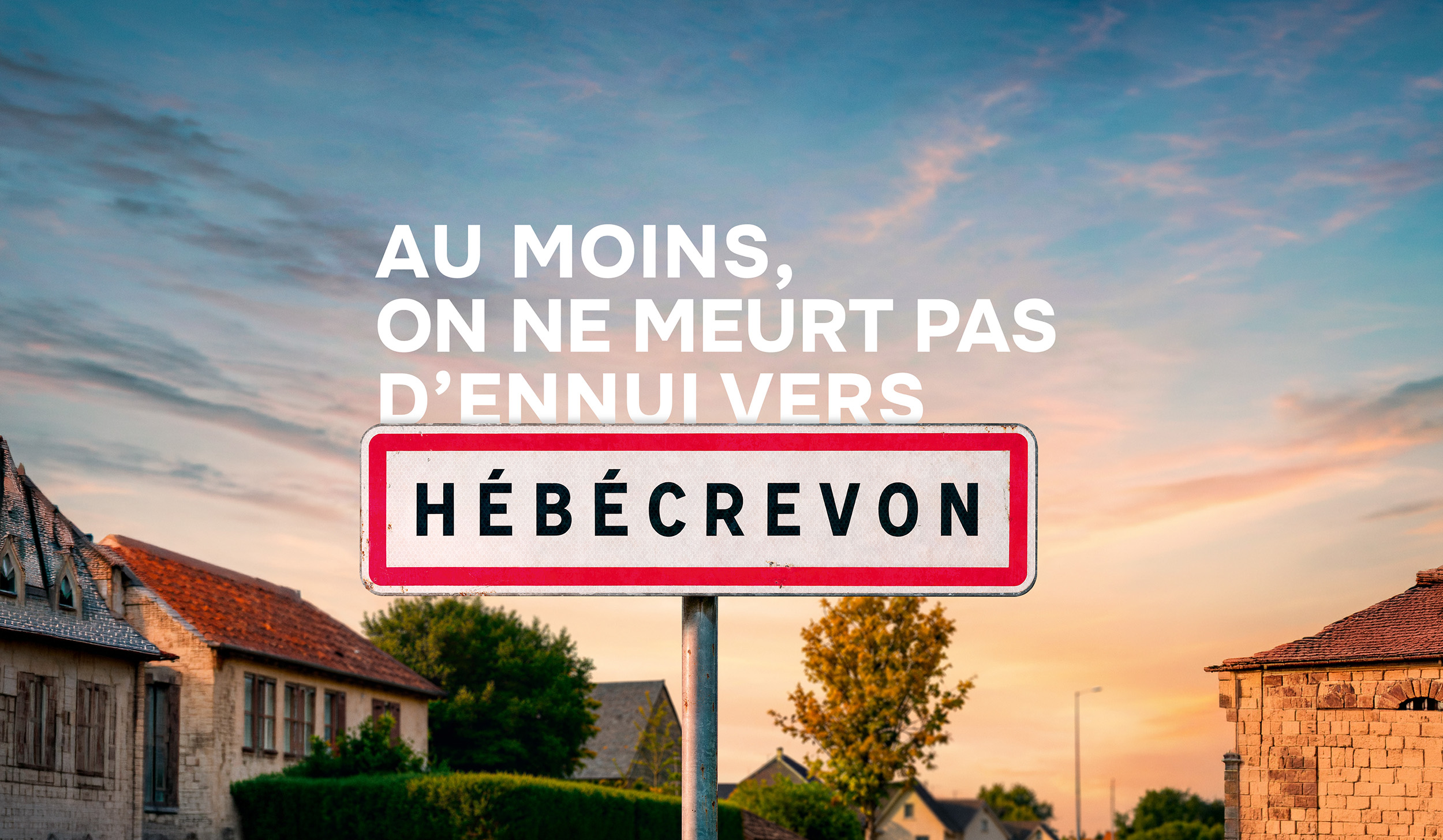 Affiche verticale JCDecaux de la nouvelle campagne PagesJaunes signée Marcel déployée à partir du 10 juillet 2026, panneau routier d'entrée d'agglomération encadré de rouge sur fond blanc portant le nom de la commune HÉBÉCREVON planté au premier plan devant un ciel de fin de journée aux nuances roses et bleues, en arrière-plan un village normand avec toits de tuiles rouges maison en pierre calcaire claire et façades chaulées, accroche « AU MOINS, ON NE MEURT PAS D'ENNUI VERS HÉBÉCREVON » en grandes capitales blanches en haut du visuel, format print vertical 120x176