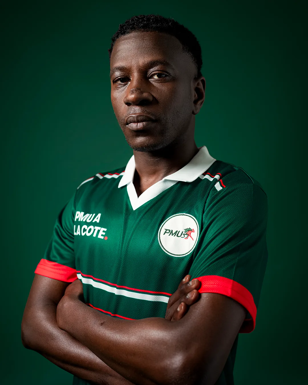 Maillot de foot signé PMU lancé le 11 juin 2026 pour la Coupe du Monde 2026, esthétique 90s vert sapin et blanc avec écusson PMU au cheval rouge brodé sur la poitrine, porté par un mannequin afro-descendant les bras croisés sur fond vert profond, photographié style sportswear pour la campagne d'activation tirage au sort