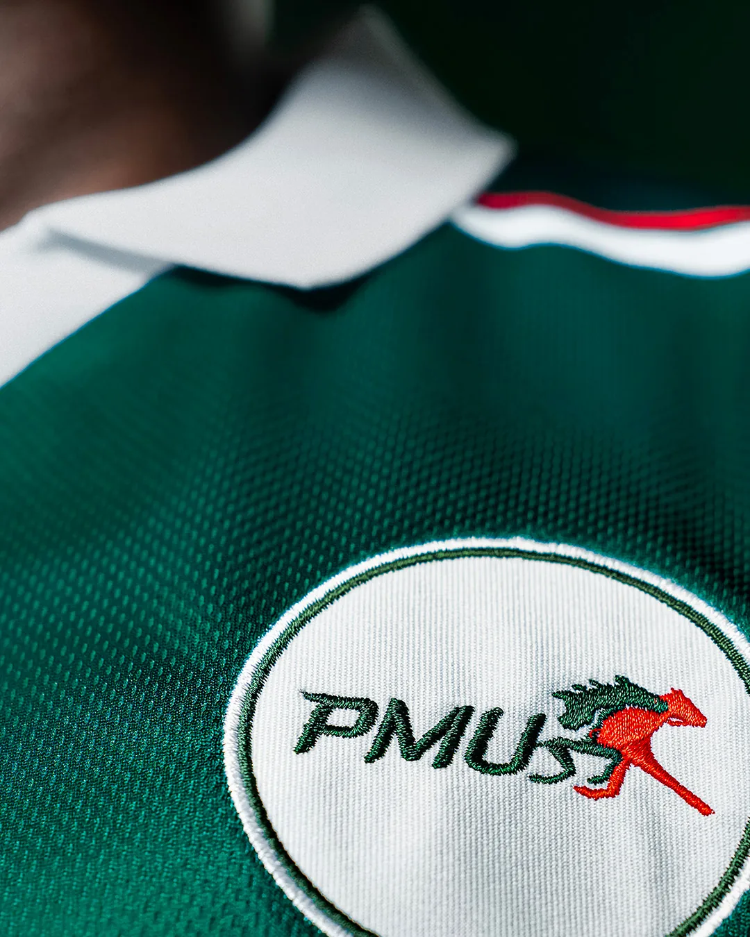 Détail brodé de l'écusson PMU sur le maillot de Coupe du Monde lancé en juin 2026, gros plan sur le blason rond blanc avec le mot PMU en lettres bouclées vert sapin et le cheval cabré rouge vermillon emblématique de la marque, encadré par le col blanc et la maille piquée vert profond du maillot type football années 90