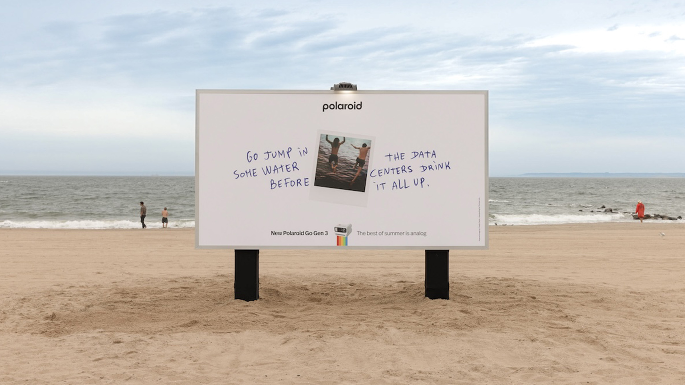 Panneau publicitaire géant Polaroid The Best of Summer Is Analog dévoilé le 19 juin 2026 planté directement dans le sable doré de la plage de Coney Island à New York, format horizontal blanc cassé avec le slogan en lettres capitales noires Go jump in some water before the data centers drink it all up, logo arc en ciel Polaroid en bas, ciel bleu pâle d'été, parasols rayés multicolores et baigneurs en arrière-plan flou