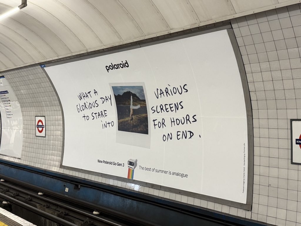 Affichage Polaroid The Best of Summer Is Analog dévoilé le 22 juin 2026 dans la station de métro King's Cross à Londres, immense panneau publicitaire au-dessus du quai à la sortie d'un train, fond crème uni avec le slogan en lettres capitales noires What a glorious day to stare into various screens for hours on end, logo arc en ciel Polaroid en bas, passagers de dos descendant du métro en silhouettes, néons des couloirs en arrière-plan flou