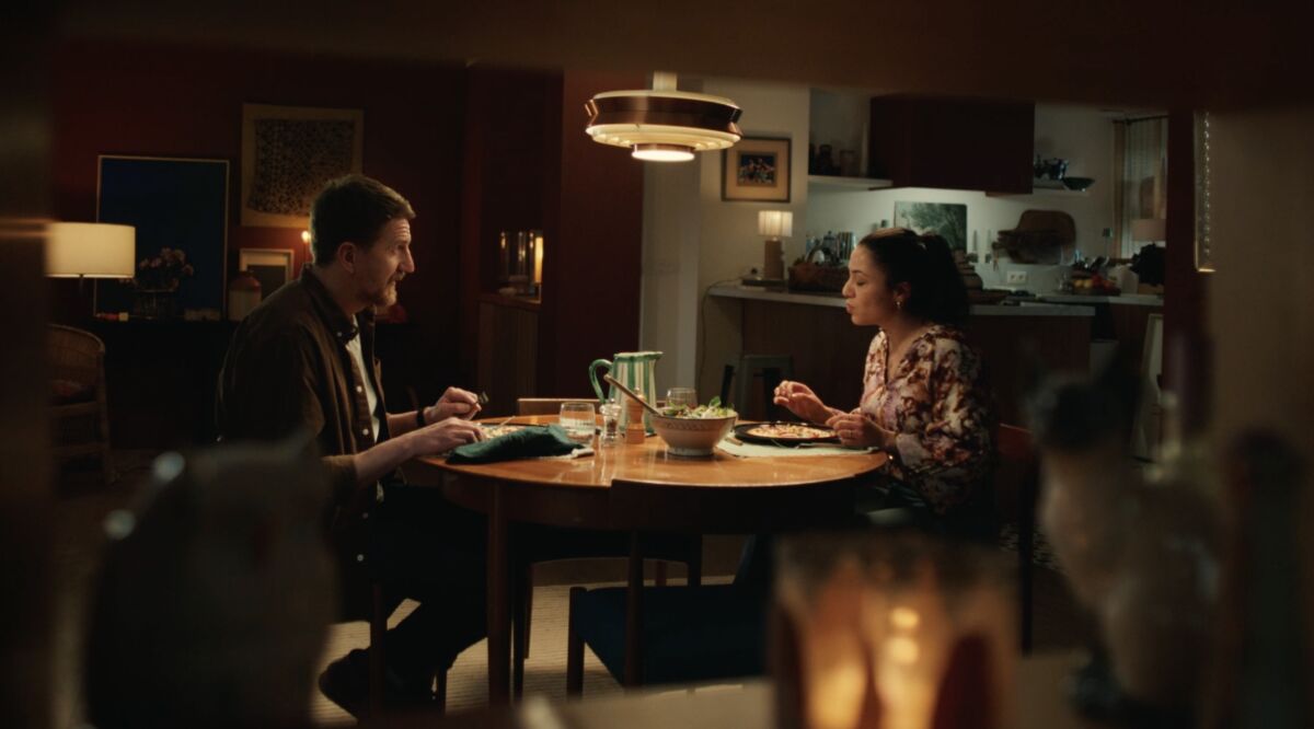 Photogramme du film 30 ans de Reflets de France signé Publicis Conseil et réalisé par Louise Condemi chez Prodigious diffusé à partir du 29 juin 2026, plan large en légère plongée dans une salle à manger cosy aux murs terracotta chauds éclairée par une grande suspension conique cuivre en fer forgé au centre, à gauche l'homme quarantaine chemise marron sur t-shirt gris barbe et cheveux châtain attablé de trois-quarts, à droite la jeune femme brune en robe fleurie prune posée face à lui, entre eux une table ronde en bois clair sur laquelle se détache une assiette bleu foncé contenant une flammekueche coupée en parts pichet en céramique verte carafe d'eau et bol de salade verte, kitchen ouverte cuisine ouverte en fond avec plans de travail bois clair et lampes de bureau blanches, décor intérieur chaleureux à l'esthétique d'auteur cinéma français