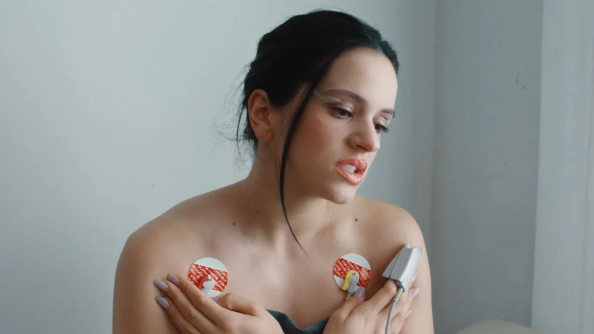 Photogramme du clip Berghain de Rosalía signé CANADÁ Barcelona récompensé du Grand Prix Entertainment for Music aux Cannes Lions 2026, plan rapproché de la chanteuse catalane cheveux noirs tirés en arrière dans une salle clinique blanche, deux électrodes circulaires blanches collées sur la poitrine reliées à un capteur médical gris tenu dans sa main droite, paupières blanches maquillées en pointe, fond gris uni
