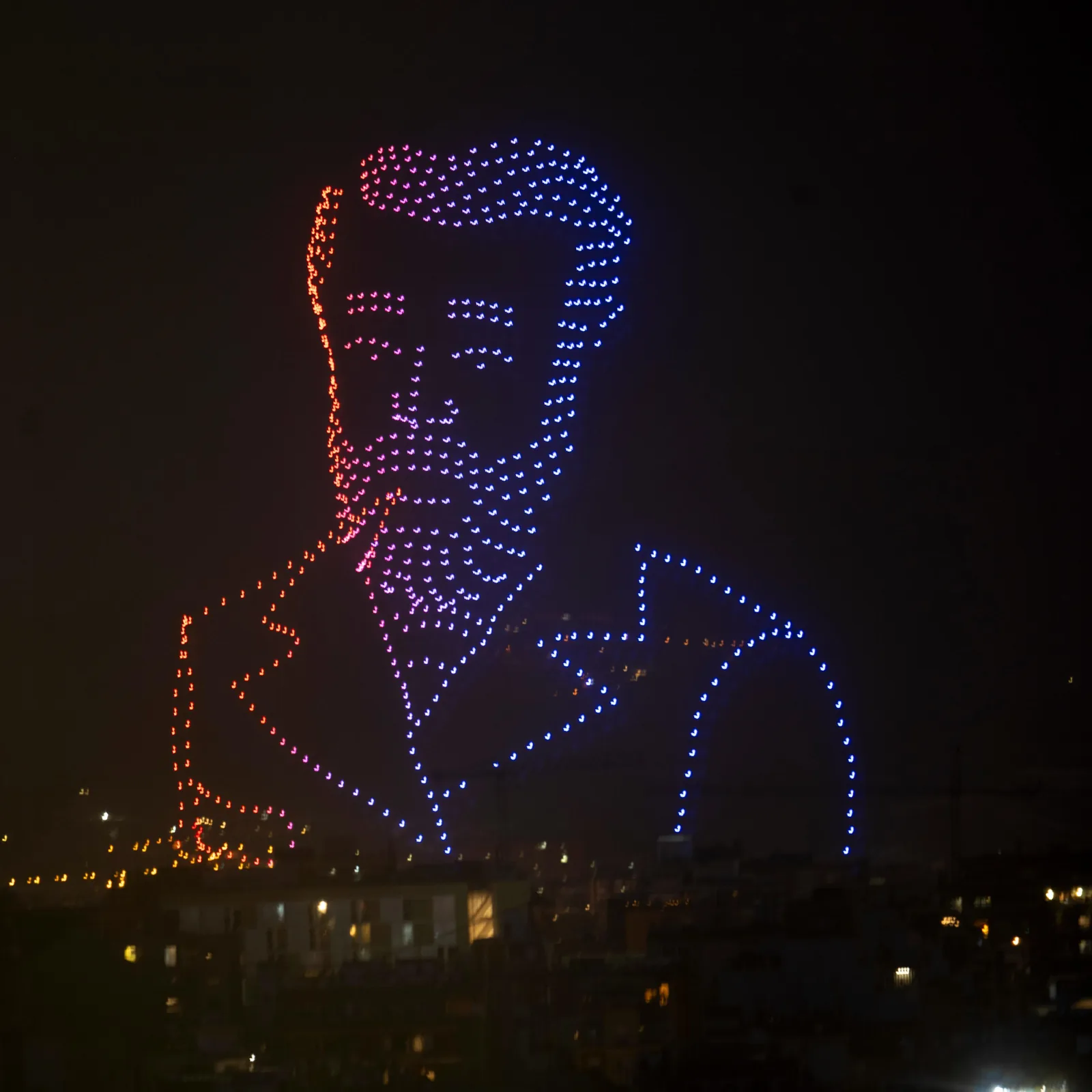 Deuxième plan du spectacle de drones du 13 juin 2026 à Barcelone, les drones forment cette fois la phrase d'Antoni Gaudí Primer l'amor, després la tècnica écrite en lumière au-dessus des flèches éclairées de la Sagrada Família, foule rassemblée sur l'avinguda Gaudí en contrebas, fumée d'ambiance bleutée flottant dans la nuit catalane