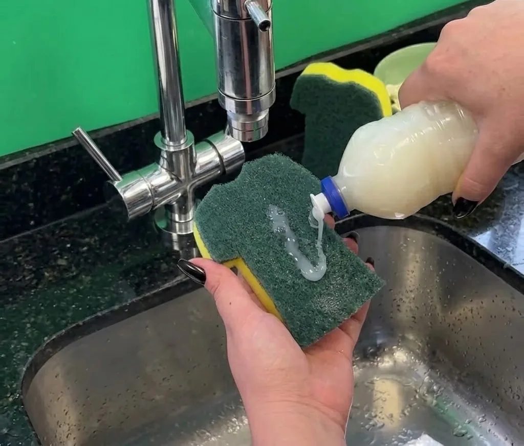 Vue de la première vague de production de l'éponge Scotch-Brite édition Coupe du Monde Brésil dévoilée le 16 juin 2026, deux éponges vert et jaune posées côte à côte sur un comptoir en bois clair d'une cuisine brésilienne avec assiettes encore sales du churrasco en arrière-plan, lumière chaude du soir, dispositif packaging dont les couleurs se devinent même sans logo