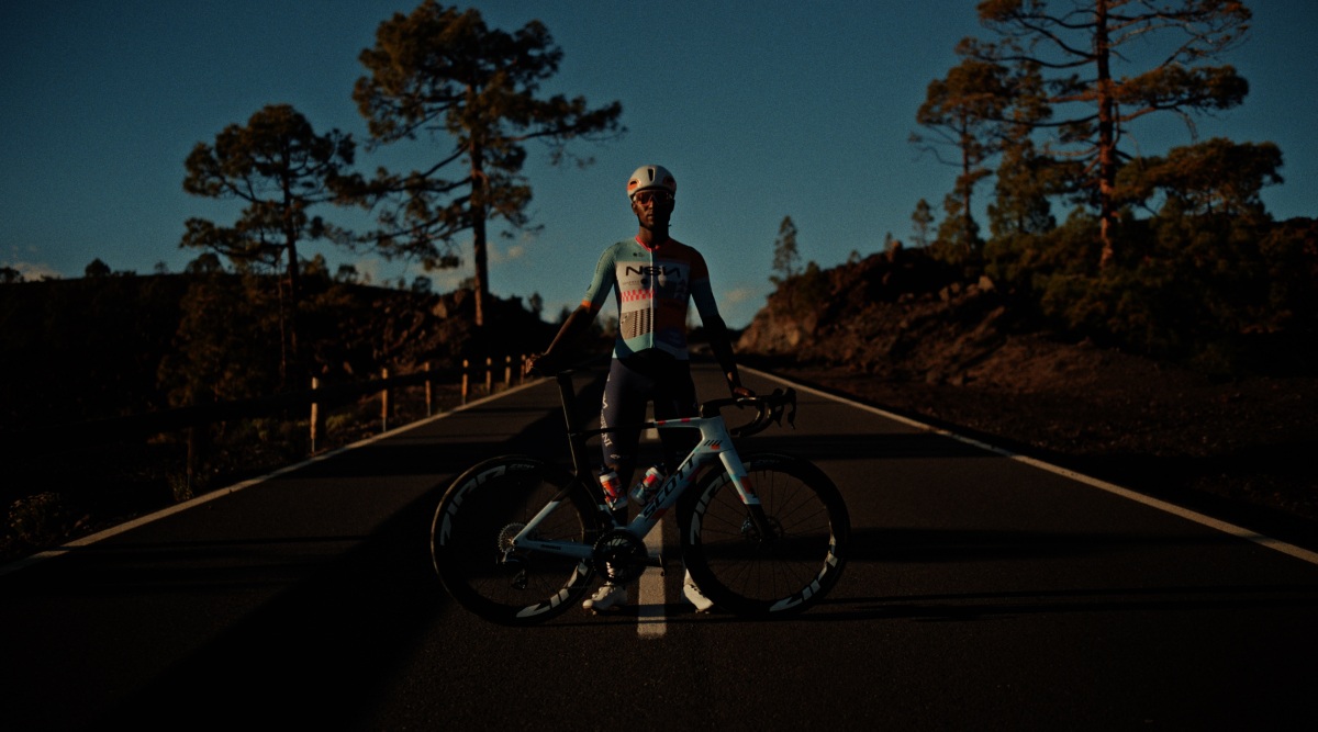 Photogramme du film manifeste SCOTT This is my Moment signé Addiction Agency avec Fifth Floor Studio et diffusé sur Eurosport pendant le Tour de France 2026, plan large frontal cinématographique en lumière rasante de fin de journée sur une route asphaltée bordée de rambardes en bois clair et de pins isolés au sommet du massif volcanique du Teide à Tenerife, ciel bleu profond dégagé sans nuage, cycliste Biniam Girmay debout au centre de la route en tenue équipe NSN maillot manches courtes bleu clair blanc et orange casque aérodynamique blanc à visière teintée main droite posée sur la selle son vélo SCOTT Foil RC blanc et bleu clair aux roues carbone Zipp posé à ses côtés, silhouette d'arbre plongée en ombre chinoise à gauche du plan, étalonnage sombre à dominante teal et orange