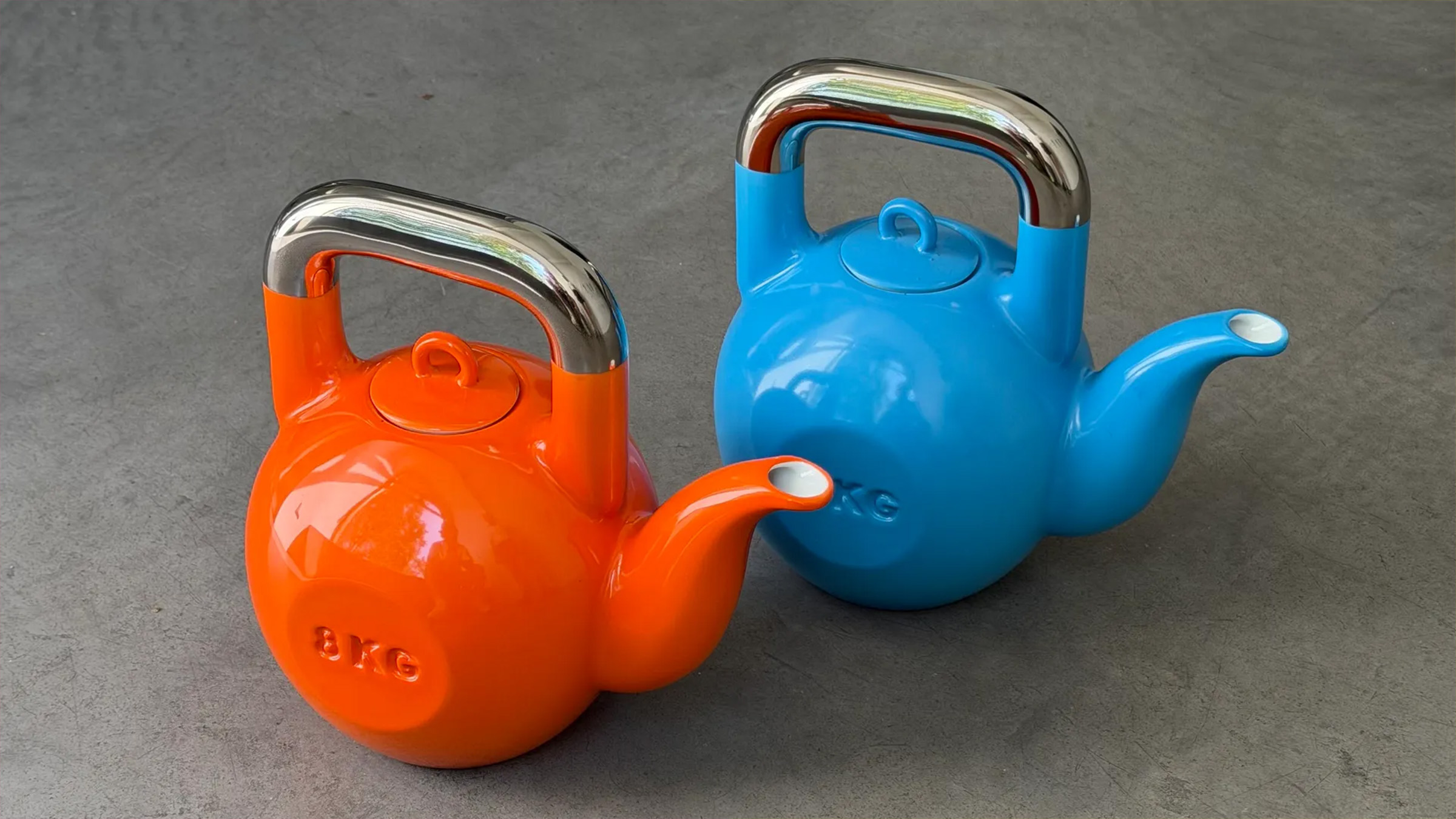 Visuel principal de la théière SporTea signée KPM Berlin et Sucuk & Bratwurst dévoilée à Catchy le 17 juin 2026, deux théières en porcelaine émaillée orange vif et bleu cobalt en forme exacte de kettlebell de musculation 8 kg avec anse rectangulaire chromée argentée brillante et bec verseur saillant peint de la même couleur que le corps, posées côte à côte de profil sur un sol béton gris foncé, mention 8 KG gravée en relief sur le flanc de chaque pièce