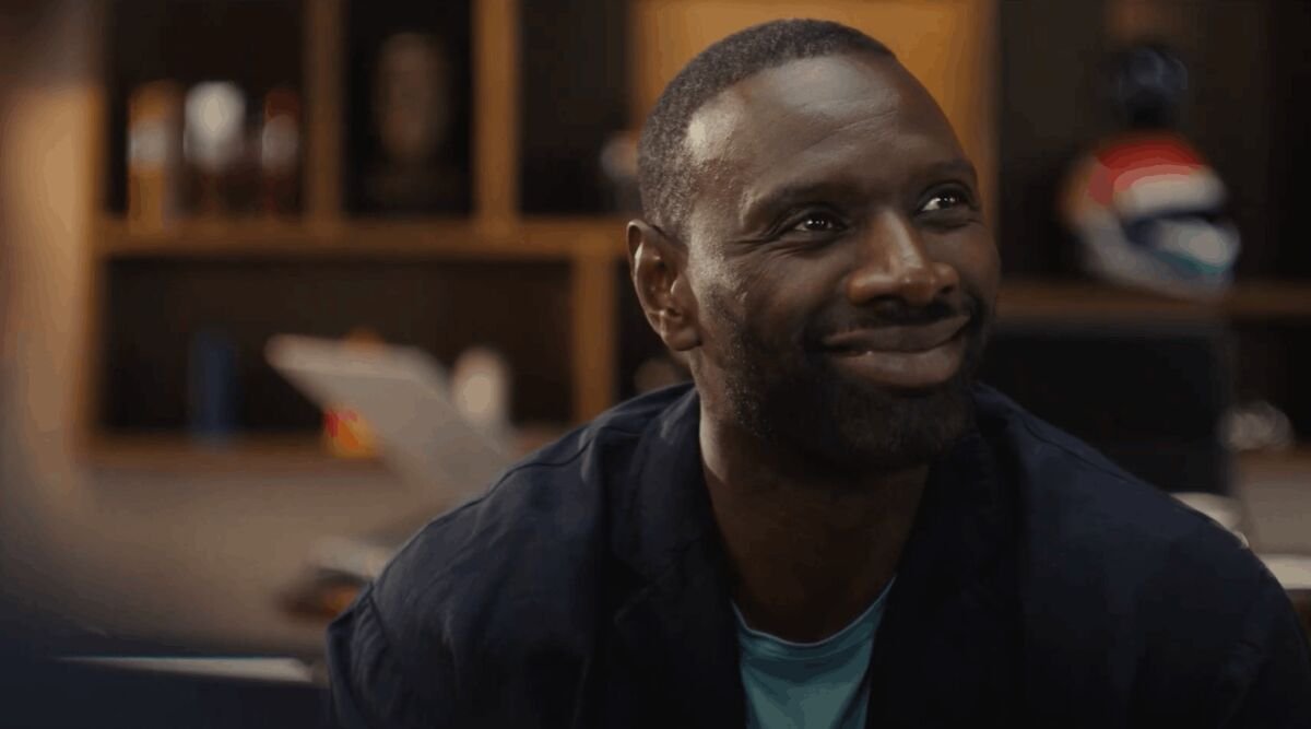 Photogramme du film Citroën devient Sytroën dévoilé le 17 juin 2026, gros plan sur le visage souriant d'Omar Sy en chemise bleu marine ouverte sur un tee-shirt turquoise, ambiance bureau intime éclairée à la lumière chaude, regard en biais et sourire complice, arrière-plan flouté avec étagères en bois sombre et touche de rouge Citroën à droite, atmosphère décontractée et populaire propre à la nouvelle signature de la marque