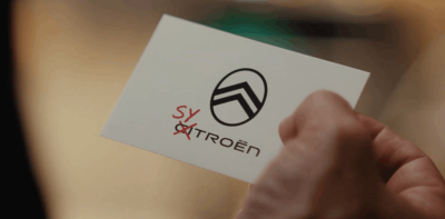 Détail du film Citroën devient Sytroën, plan rapproché sur une main qui tient le badge Citroën blanc avec le nouveau lettrage Sytroën en rouge, les lettres SY sont rajoutées à la main par-dessus le sigle officiel comme une rature manuscrite, fond flouté chaud aux teintes ocre et orangé, photo prise au moment où Omar Sy rebaptise symboliquement la marque dans la séquence finale du spot