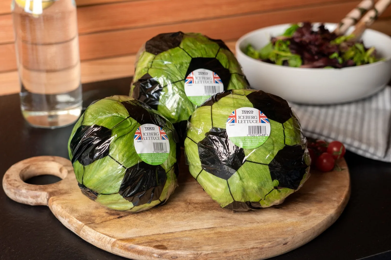 Mise en scène des laitues iceberg Tesco emballées en ballon de foot pour la Coupe du Monde 2026, trois laitues posées sur une planche à découper en bois, chacune recouverte d'un film plastique imprimé en damier hexagonal noir et blanc imitant la peau d'un ballon de foot classique, étiquette ronde Tesco Iceberg Lettuce drapeau britannique visible, bol de salade verte et tomates cerises en arrière-plan
