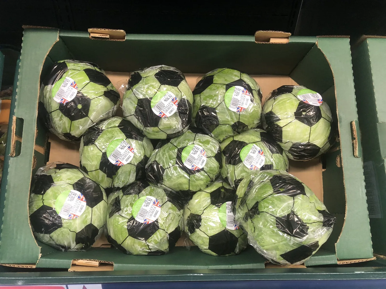 Vue plongeante d'un casier d'entrepôt Tesco contenant la première vague de laitues iceberg ballon de foot livrée le 15 juin 2026 pour la Coupe du Monde, douze laitues alignées dans une caisse en carton vert frappée du logo Tesco, chacune emballée du film plastique damier noir et blanc, étiquettes rouges à drapeau britannique visibles sur chaque emballage