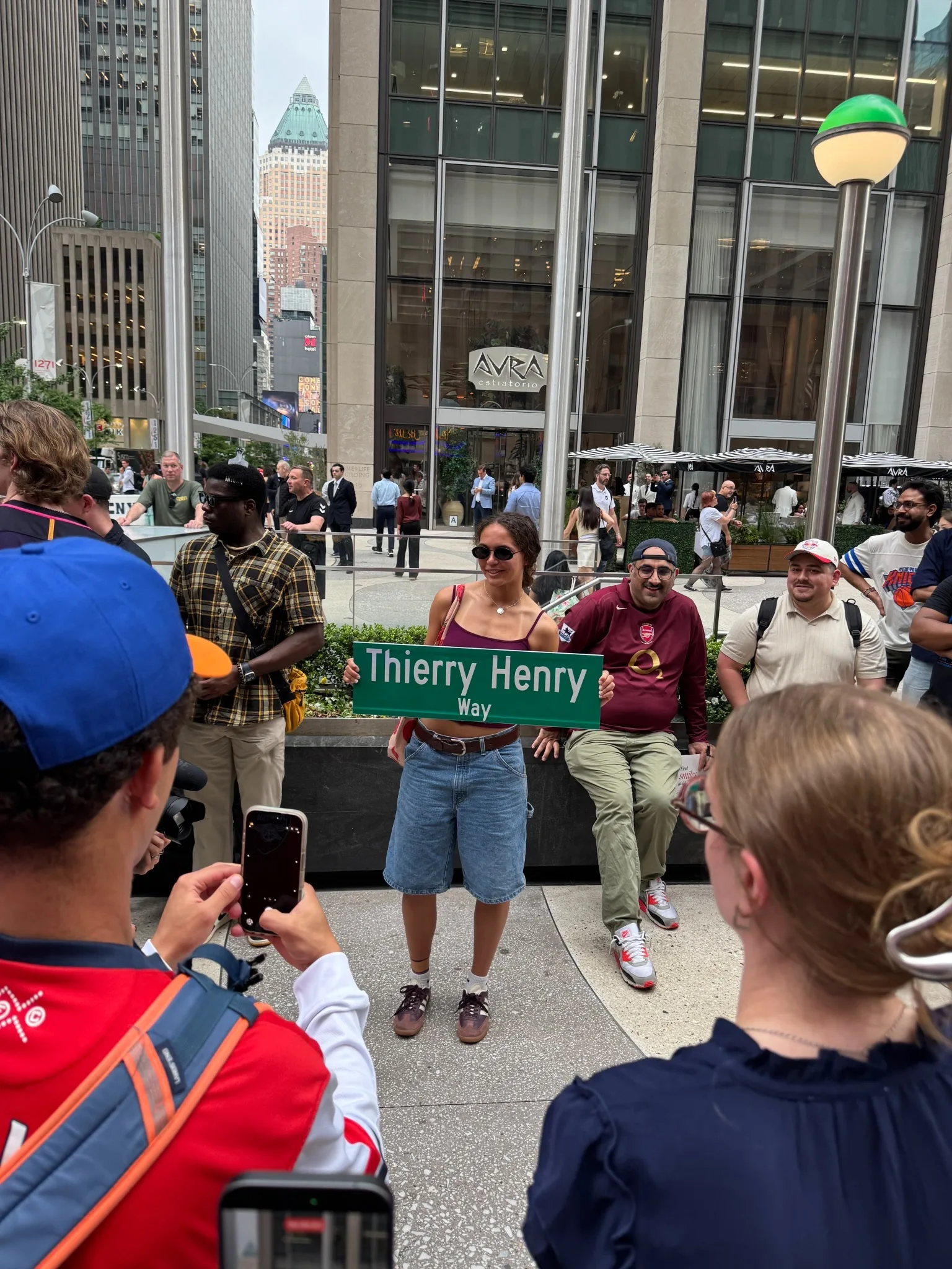 Photographie de la cérémonie d'inauguration de la Thierry Henry Way le 12 juin 2026 à Manhattan, la conseillère municipale Virginia Maloney présente le nouveau panneau bleu et blanc devant un parterre de supporters et de journalistes, façade en briques rouges typique du Midtown à l'arrière-plan, cérémonie officielle de la ville de New York