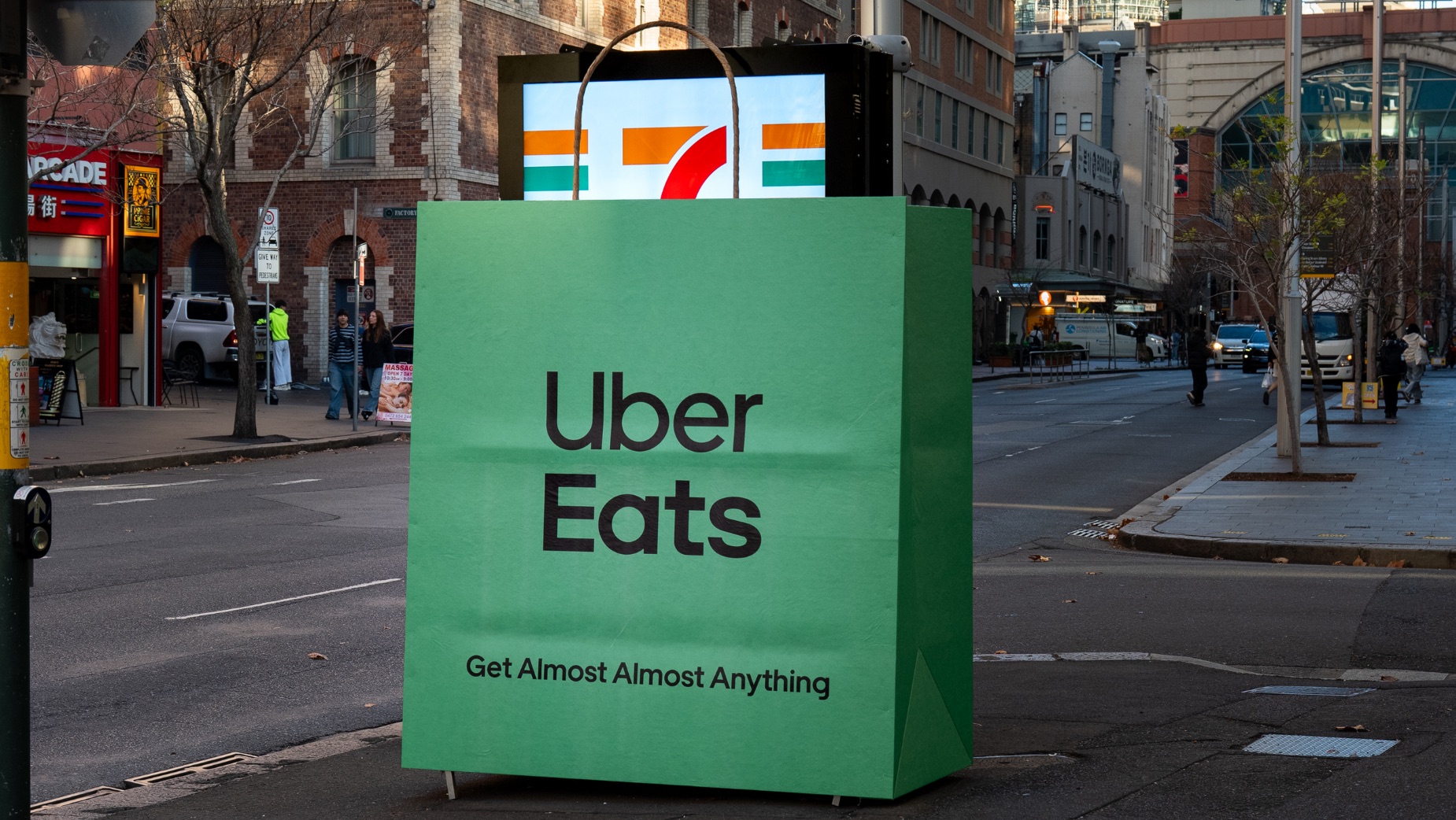 Special build Uber Eats Australie dévoilé le 18 juin 2026 sur un panneau publicitaire vu en plan rapproché, sac de livraison vert sapin frappé du logo Uber Eats en blanc recouvrant partiellement l'affiche McDonald's, sangle visible nouée en surimpression du panneau, environnement de banlieue australienne avec ciel pâle et bâtiments en briques, photographie en lumière naturelle de jour