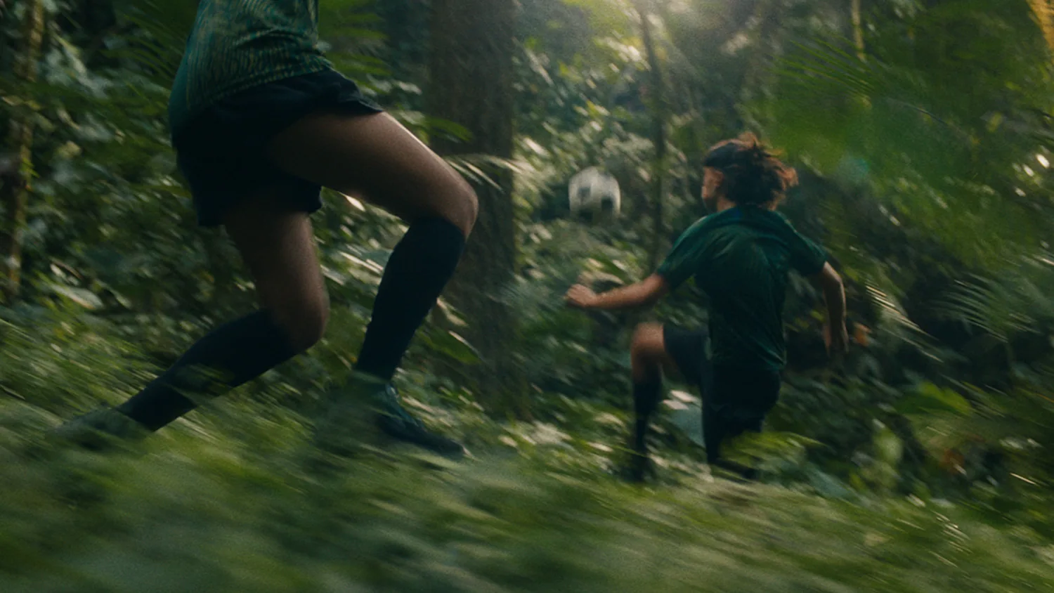 Capture du film Team Forest signé WWF et Studio Birthplace dévoilé le 11 juin 2026, deux jeunes footballeurs courent en plein effort dans la forêt tropicale d'Oxygène à Sumatra avec ballon blanc en arrière-plan, motion-blur et lumière dorée traversant la canopée, équipement aux couleurs de la forêt vert sombre et noir