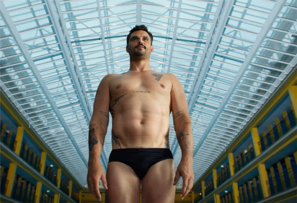 Photogramme extrait du film Le Ballet réalisé pour Yomoni par l'agence Brainsonic dévoilé le 4 juillet 2026, plan en contre-plongée américaine du champion olympique de natation Florent Manaudou en maillot de bain noir moulant style boxer debout jambes légèrement écartées bras le long du corps regard porté vers le haut vers l'immense verrière en arc de cercle vitrée blanche caractéristique de la piscine Molitor à Paris, tatouages tribaux visibles sur son épaule gauche et son bras droit texte tatoué sur la poitrine, corps musclé bronzé cheveux courts châtain et moustache brune, en arrière-plan la structure métallique blanche en berceau de la verrière et les alignements jaune vif des cabines individuelles du bassin d'hiver, tirage cinéma haute définition étalonnage doux légèrement bleuté