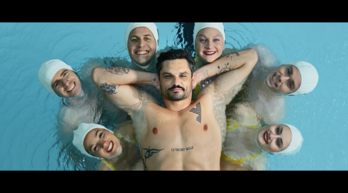 Photogramme du film « Le Ballet » de Yomoni signé Brainsonic et réalisé par ARGO, plan aérien vertical en plongée sur la surface d'un bassin de la piscine Molitor à Paris, Florent Manaudou allongé au centre sur le dos les yeux fermés torse nu à peau claire mains croisées derrière la nuque tatouages tribaux et inscription La vie est belle sur le buste, six membres de l'équipe de France de nage synchronisée disposés autour de lui en couronne symétrique tête en avant bonnets de bain blancs souriants deux à droite deux à gauche et deux en haut de la formation, eau bleu turquoise miroitante avec reflets clairs, cadrage cinématographique bord noir en haut et en bas format cinéma