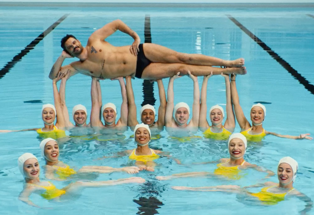 Photogramme du film publicitaire Le Ballet de Yomoni tourné par Brainsonic à la piscine Molitor à Paris pour un lancement le 4 juillet 2026, plan large en plongée légère au-dessus du grand bassin d'eau turquoise claire, le champion olympique Florent Manaudou allongé sur le côté en maillot noir la tête posée sur son bras plié et les jambes tendues levé horizontalement au-dessus de la surface porté à bout de bras par une figure de nage synchronisée impeccable des membres de l'équipe de France Olympique, douze nageurs et nageuses en bonnets de bain blancs et maillots jaunes canari immergés jusqu'à la poitrine forment une pyramide humaine en U autour de lui grands sourires face caméra bras tendus et jambes ancrées sous l'eau, lignes de nage noires visibles sous la surface transparente, éclairage doux et étalonnage cinéma
