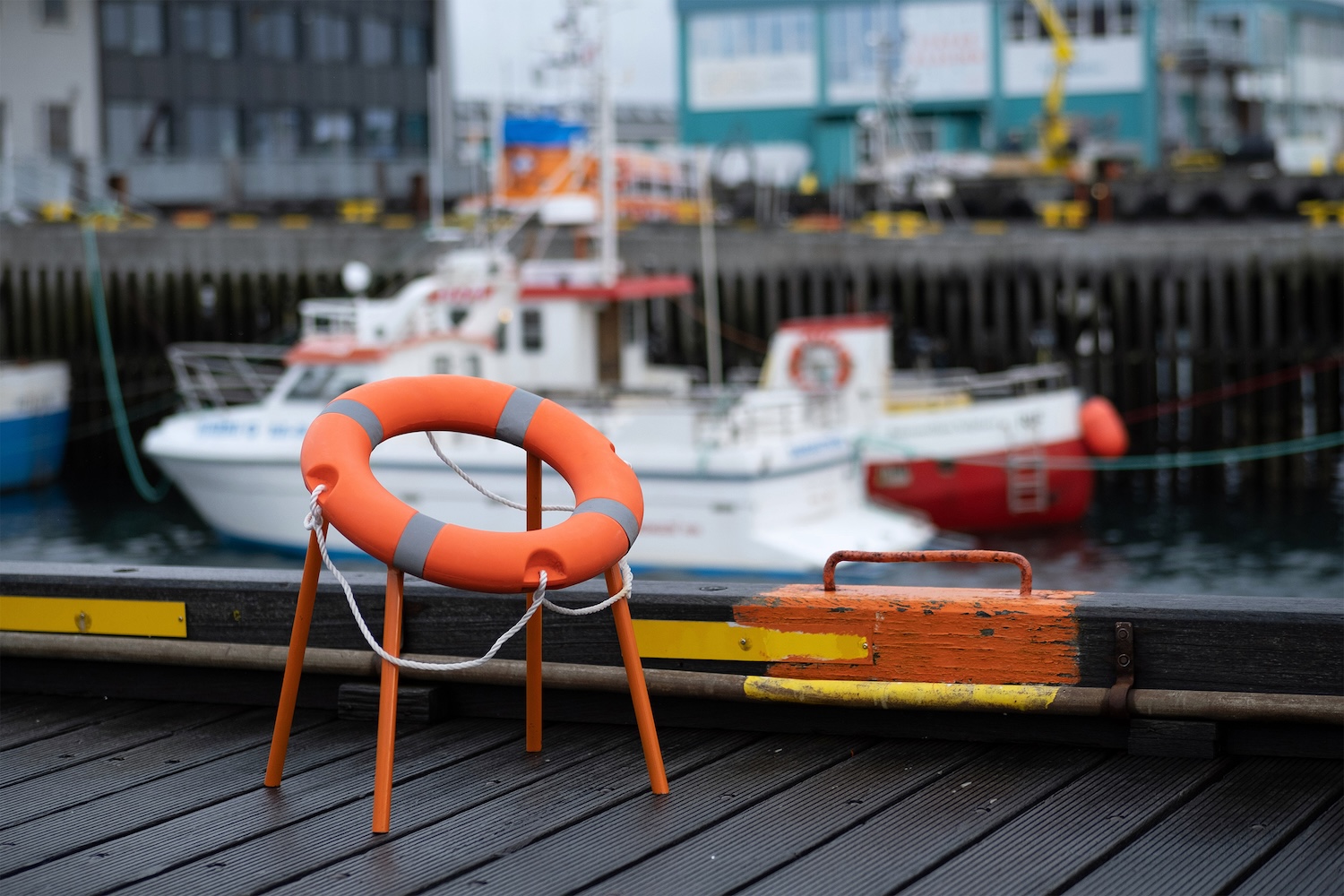 Photographie diurne en plan rapproché horizontal de la Sea Level Rise Chair de Tobia Zambotti posée sur un ponton en bois foncé à Reykjavík en juillet 2026, chaise structure métallique fine orange vif à quatre pieds inclinés supportant une bouée de sauvetage circulaire orange à bandes réfléchissantes grises et corde blanche tressée qui pend, au premier plan à droite un bollard en bois foncé peint d'orange écaillé fixé par une pièce métallique rouillée, arrière-plan flou d'un port d'Islande avec deux bateaux de pêche blancs à coque rouge alignés le long du quai et devanture d'un hangar portuaire vert turquoise à plusieurs fenêtres, sol du quai en lattes de bois foncées et bandes latérales peintes en jaune et rouge, atmosphère grise et humide de matin océanique nordique